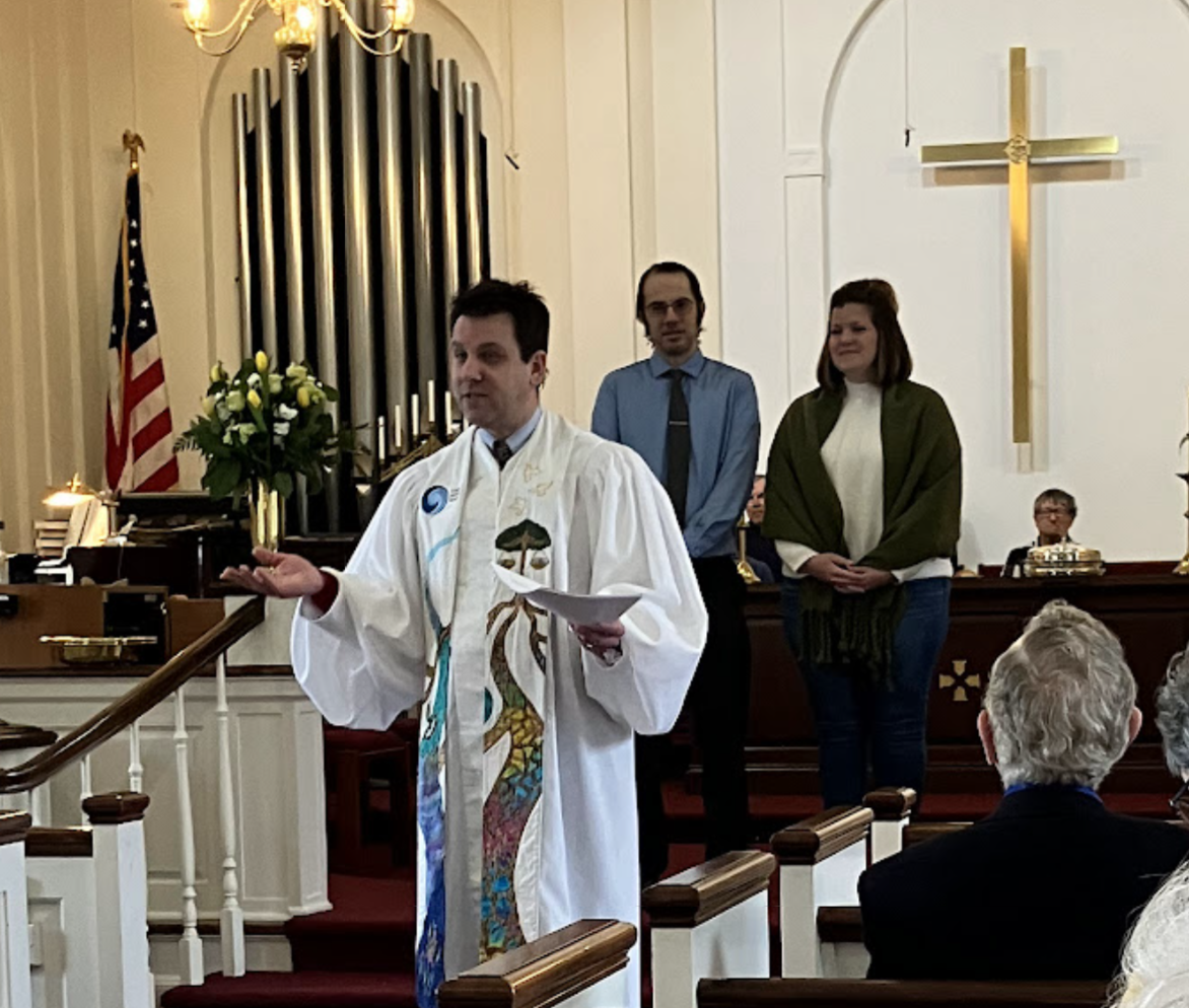 A pastor in ceremonial robes conducting a service inside a church with a cross on the wall and an American flag. Two people stand behind him, and a woman wears a green shawl. Several congregants are seated, listening attentively.