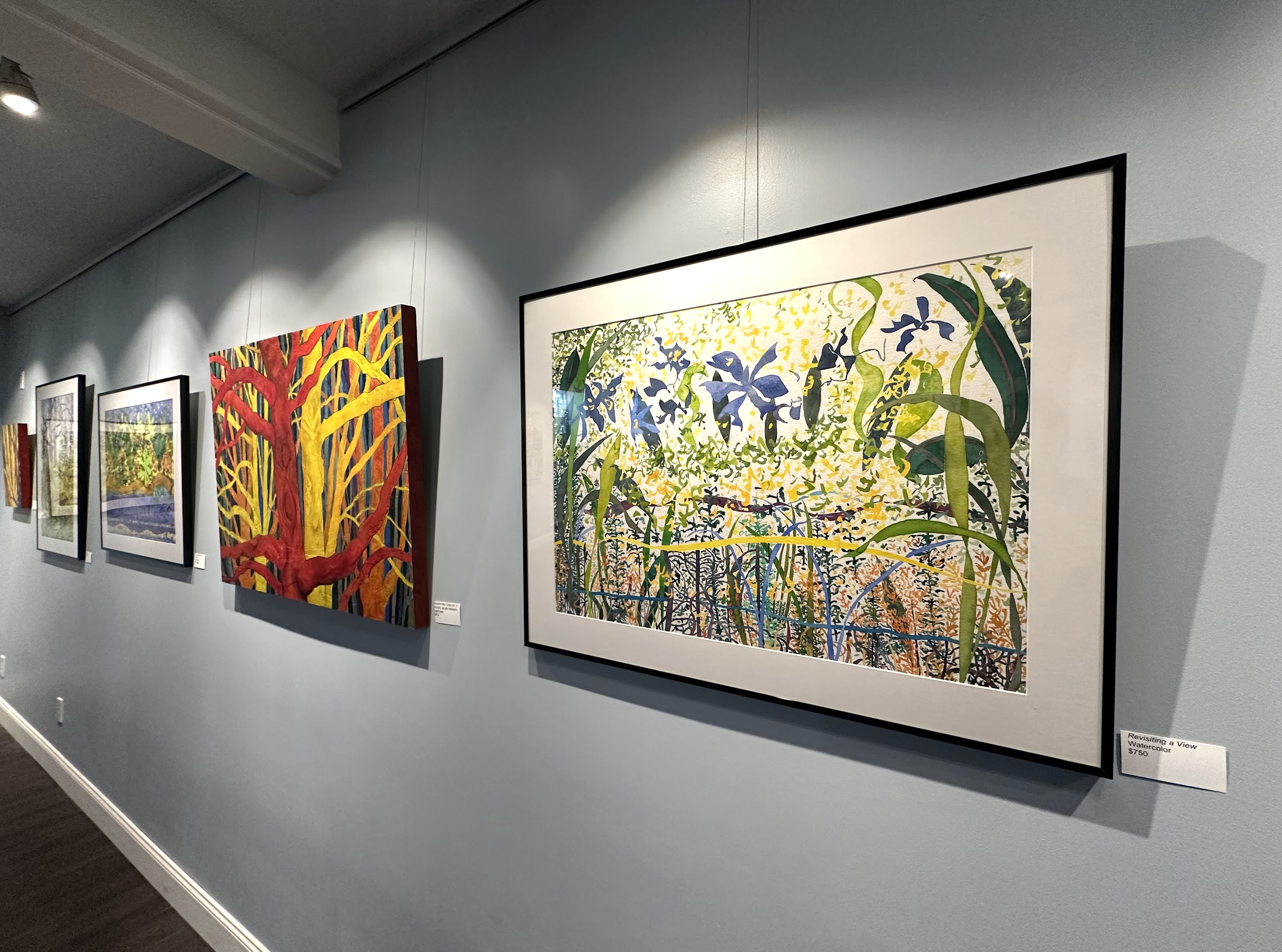 An art gallery wall displaying colorful paintings, including a large vibrant depiction of a dense forest with red and yellow trees and a lush landscape with flowers and plants.