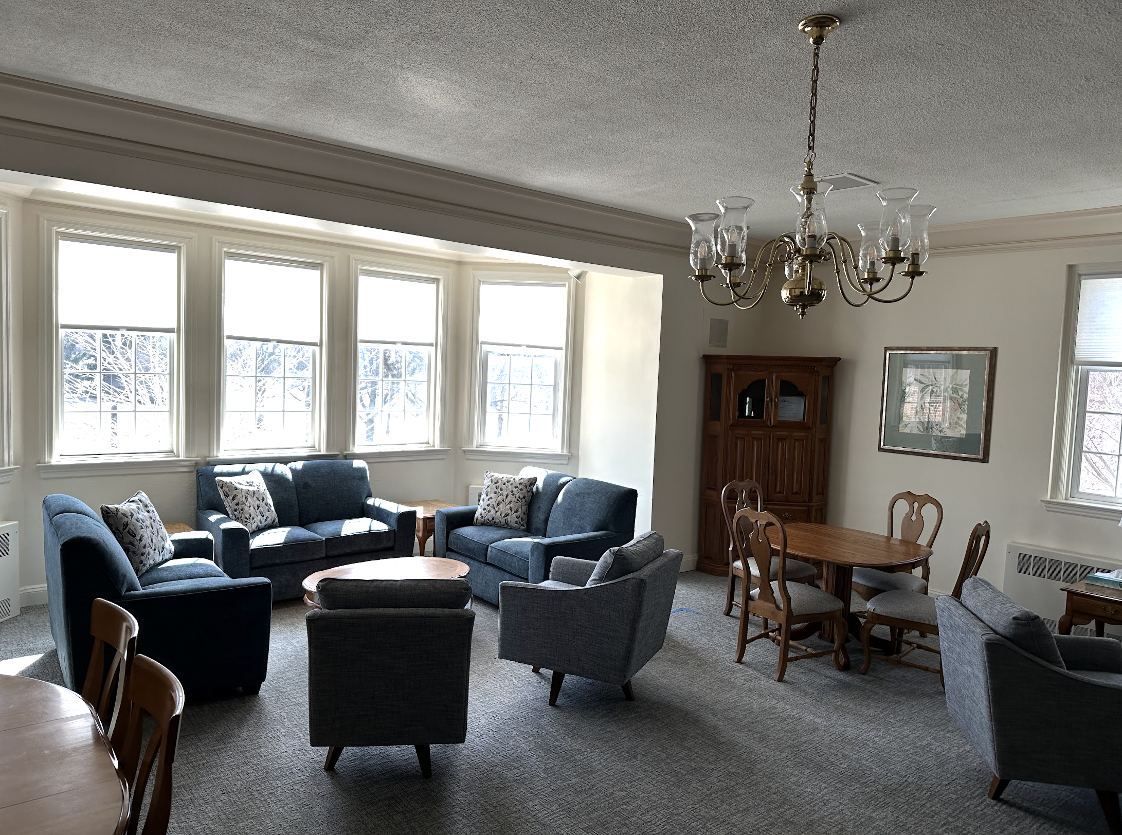 The church lounge room with a large bay window, blue sofa set with patterned pillows, four gray armchairs, a wooden dining table with chairs, a wooden cabinet, framed picture on the wall, chandelier hanging from the ceiling, and carpeted floor.