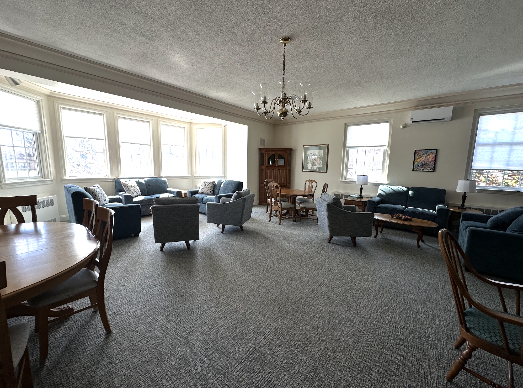 The church lounge with multiple blue sofas and gray armchairs arranged around coffee tables, natural light from large bay windows, carpeted floor, chandelier, framed artwork on walls, and wooden dining table with chairs.