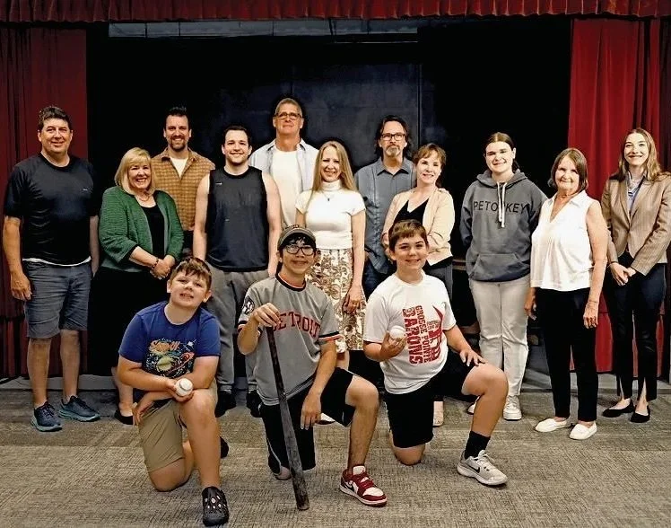 Group of people posing for a photo in front of a stage with red curtains, including adults and children, some smiling and others with serious expressions.