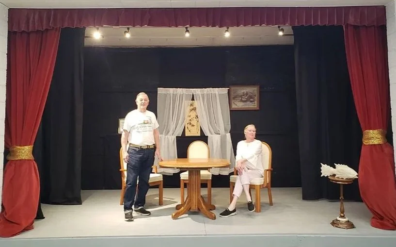 A man standing and a woman sitting on stage in a small theater or play setting with red curtains, black backdrop, table, two chairs, window with curtains, and wall art. Theater performance. 
