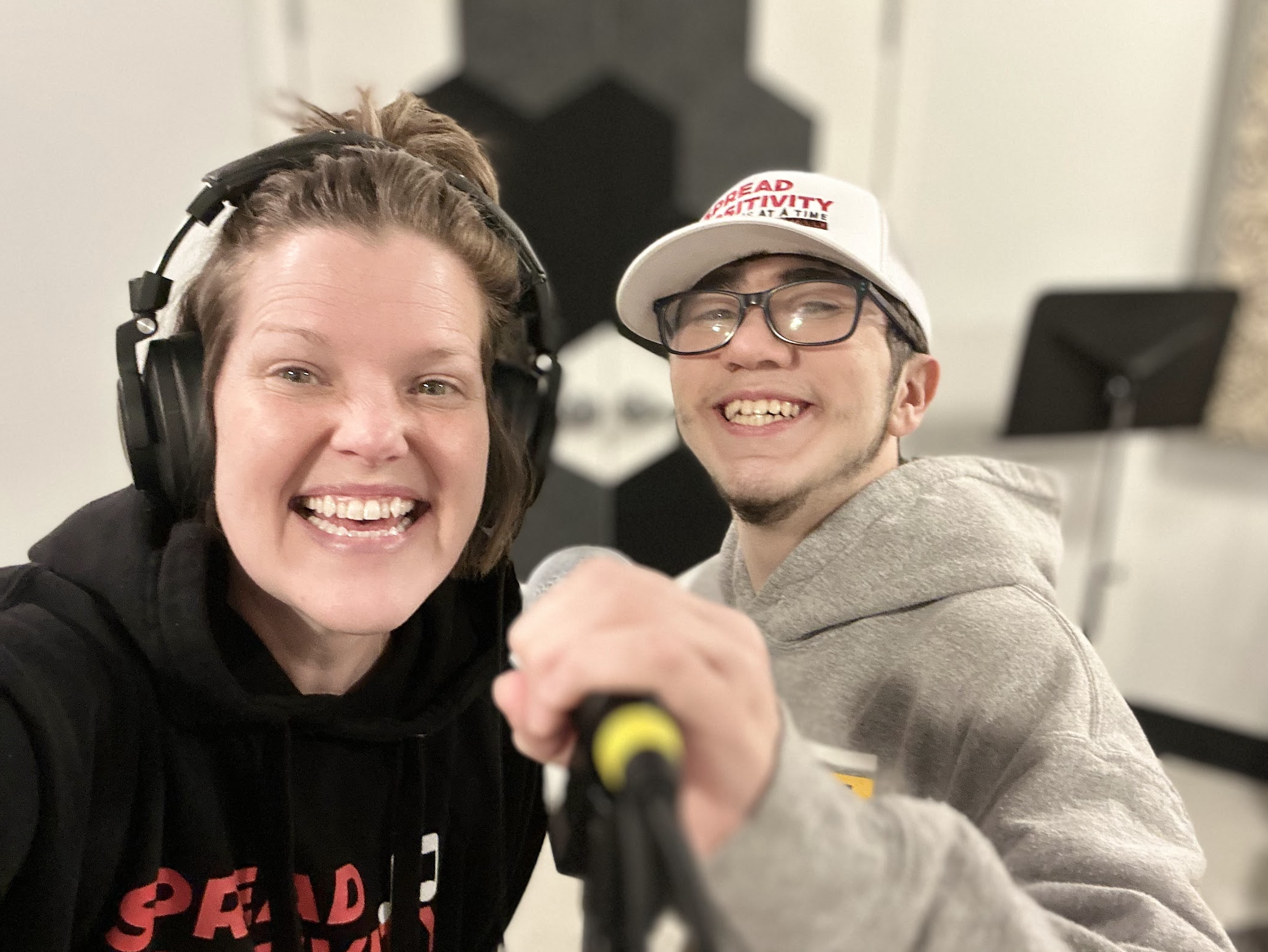 Two people smiling, one wearing large headphones and the other holding a microphone, in a music studio.