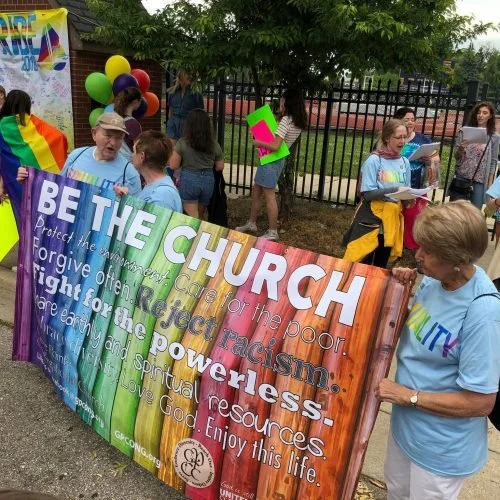 People gathered at an outdoor LGBTQ+ pride event, holding a colorful banner that promotes inclusivity, anti-racism, and love. Some individuals wear rainbow-themed clothing, and balloons are visible in the background.