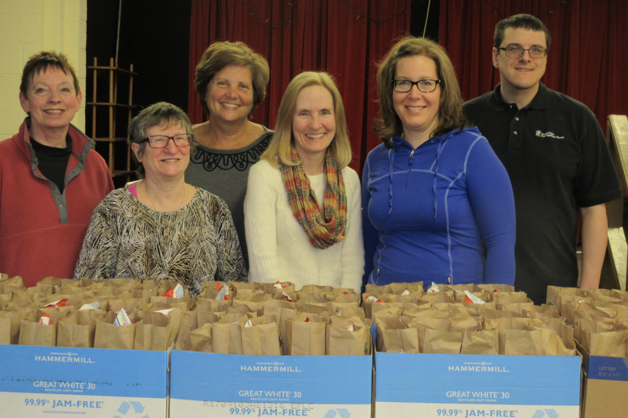 Group of seven smiling people standing behind boxes of paper bags on a table, with a red curtain and stage in the background.