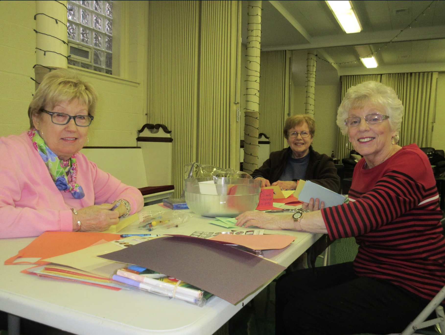 Three older women sitting at a table with colorful paper and crafting supplies, smiling in a decorated room.