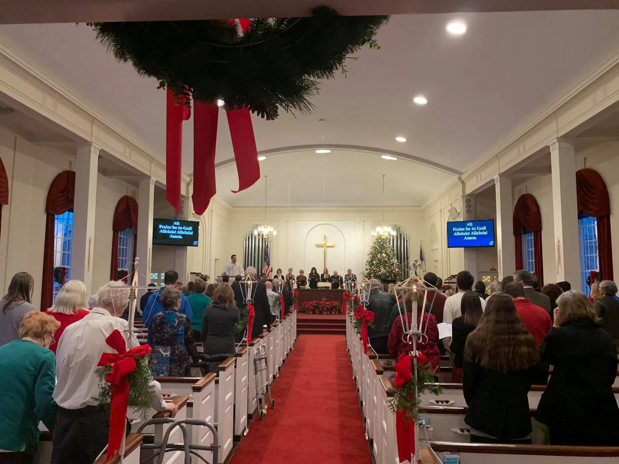 A church decorated for Christmas with a congregation standing during a service. There is a large Christmas tree, a cross on the wall, and red bows with holiday decorations on the pew railings.