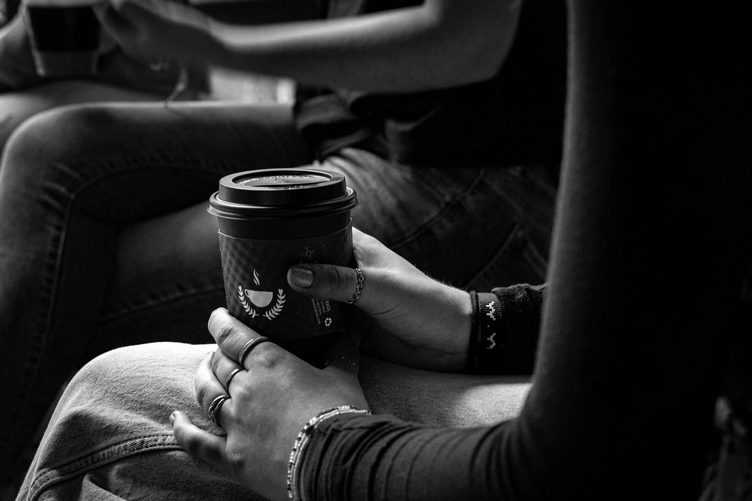 Close-up of a person holding a to-go coffee cup with both hands, seated with legs crossed, wearing rings and bracelets, in a monochrome setting.