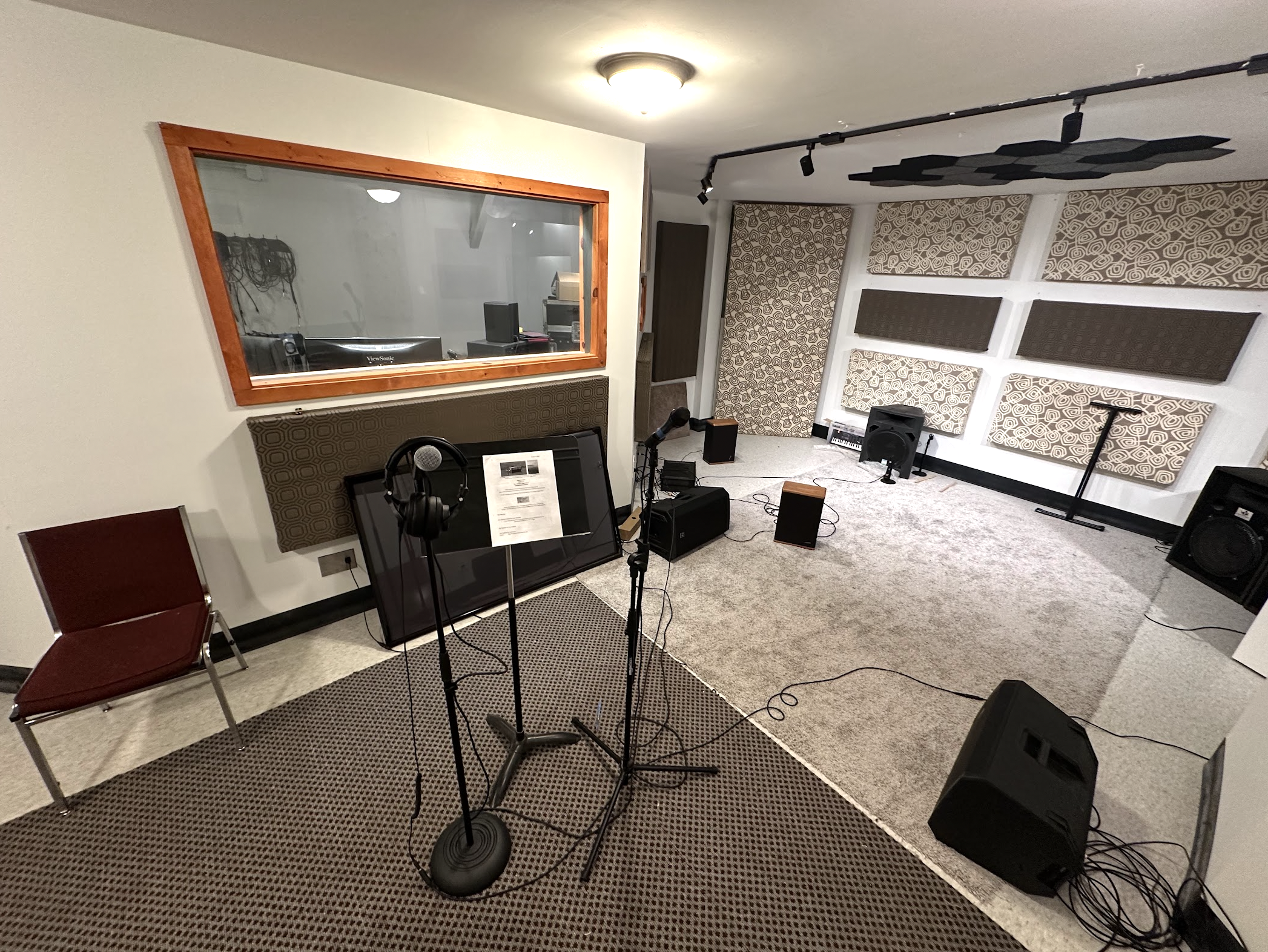 Recording studio with a microphone stand, headphones, and music equipment on the floor, a window looking into another room, acoustic panels on the walls, and a chair to the left.