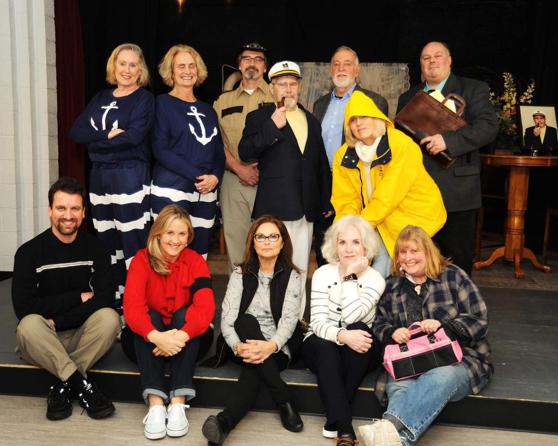 A group of 12 people posing together on a stage, with some dressed in nautical-themed costumes and others in casual attire, in front of a black background and stage props.