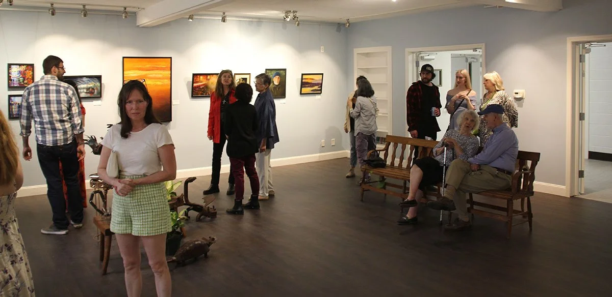 People viewing artwork in an art gallery, with paintings hung on the wall and visitors engaging with each other.