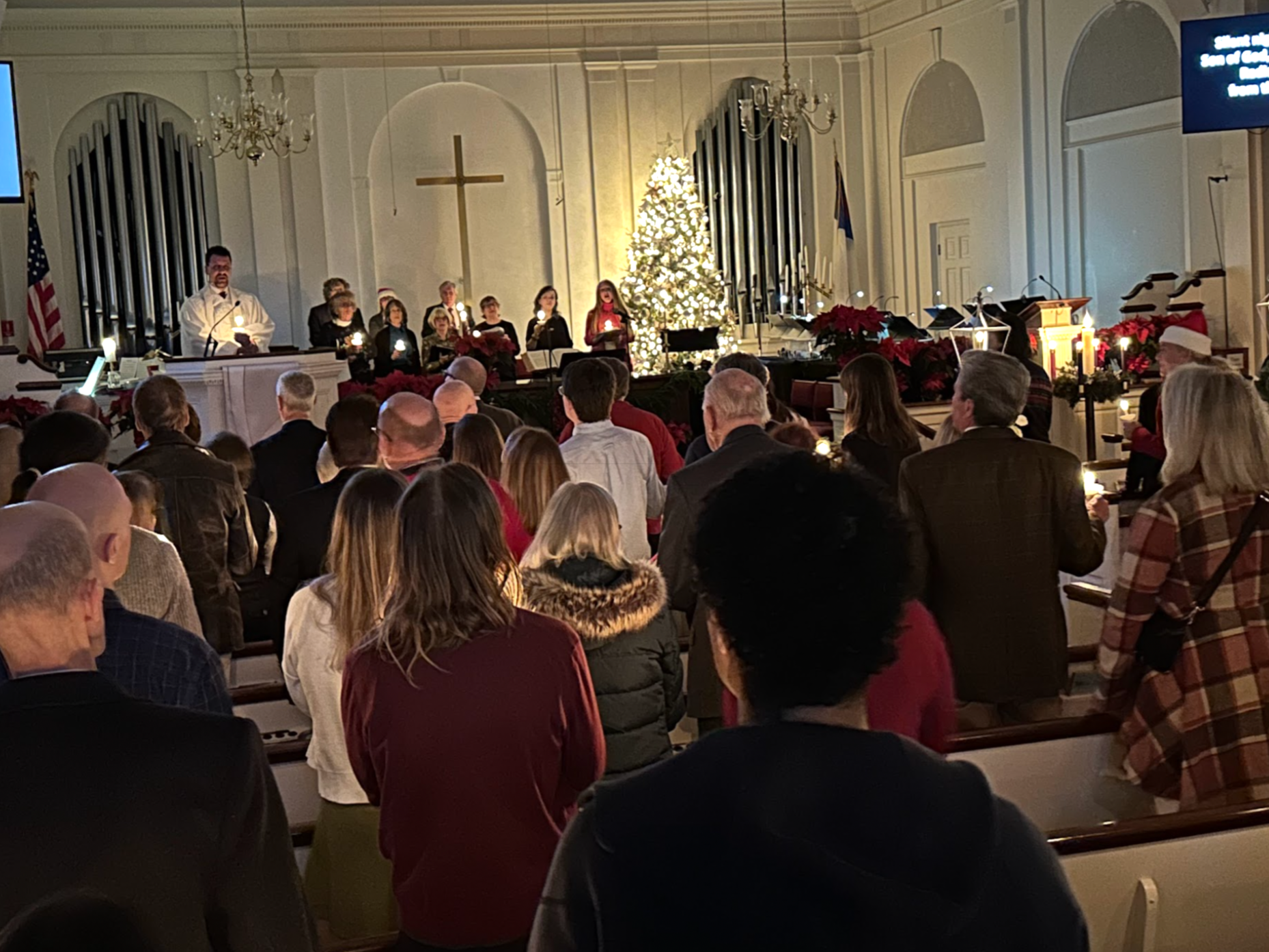 A Christmas service inside a church, decorated with a Christmas tree, poinsettias, and candles, with a choir singing in the background and congregants seated in pews.