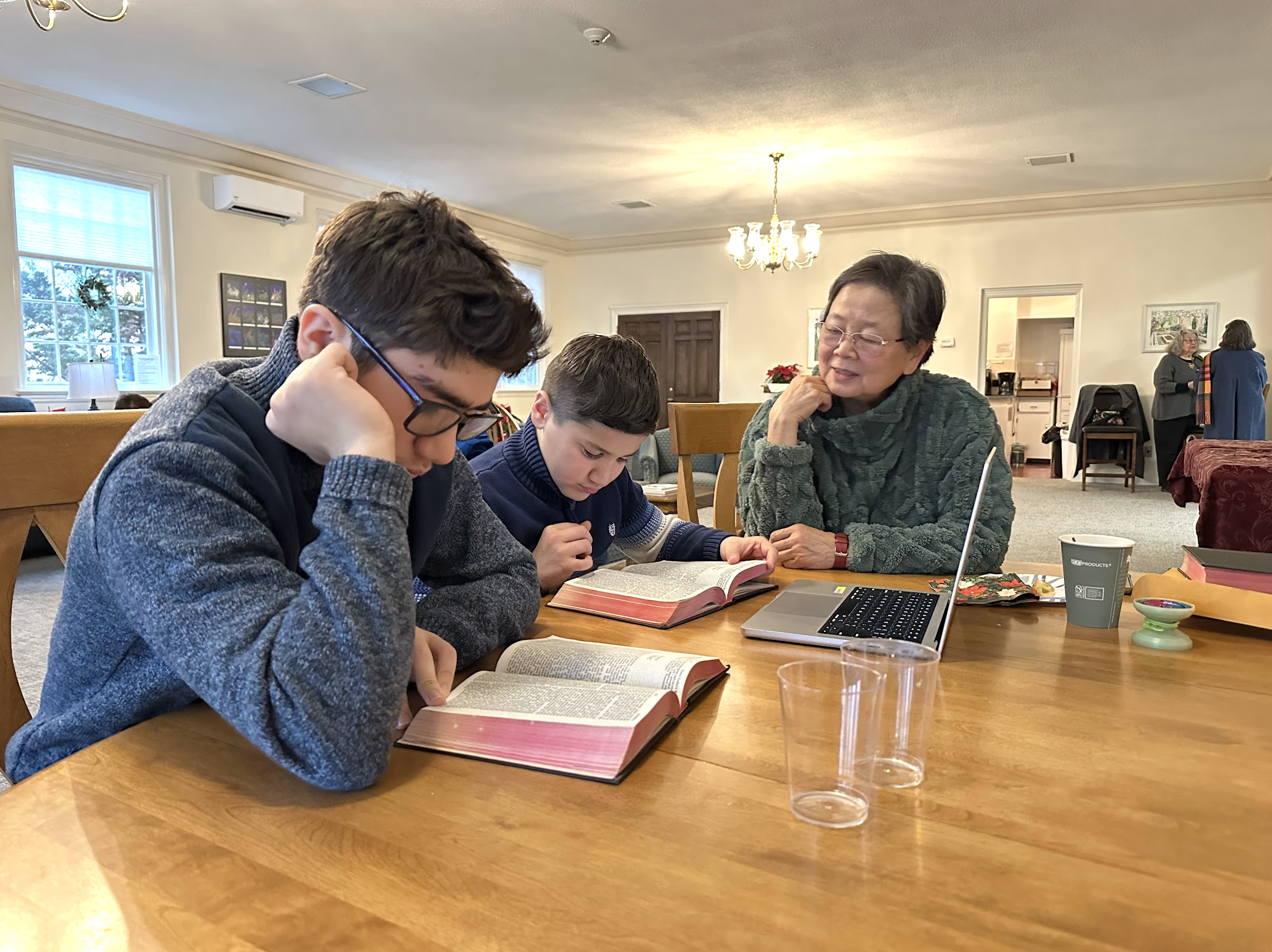 Three people, two children and an older woman, sitting at a wooden table reading from large books, in a spacious church lounge room with windows and a chandelier.