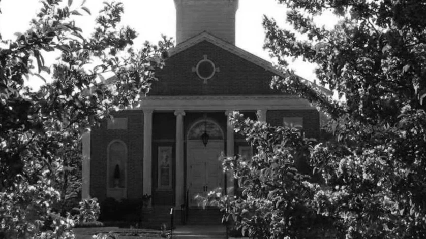 Black and white photo of a church with a prominent entrance, columns, and a bell tower, surrounded by trees and bushes.
