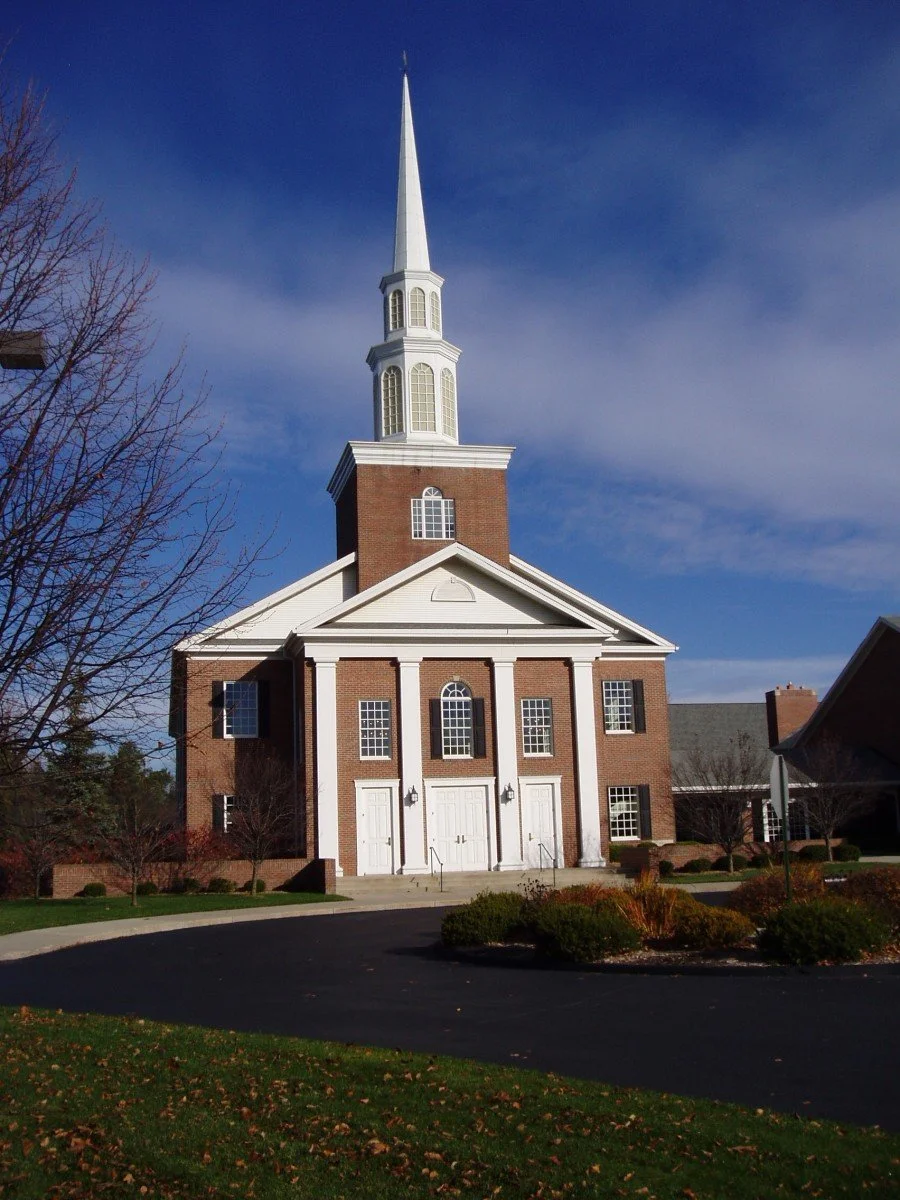 A church with red brick walls, white columns, black shutters, and a tall white steeple against a blue sky.