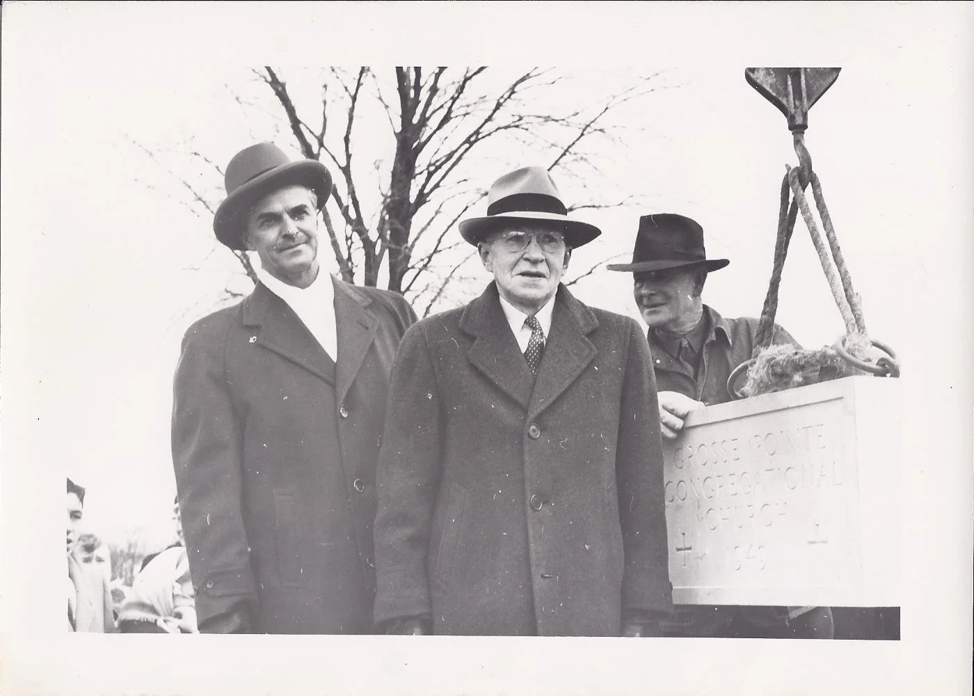 Black and white photo of three men standing outdoors, all wearing hats and coats, with a leafless tree in the background. The man on the right is holding a lever connected to a box.