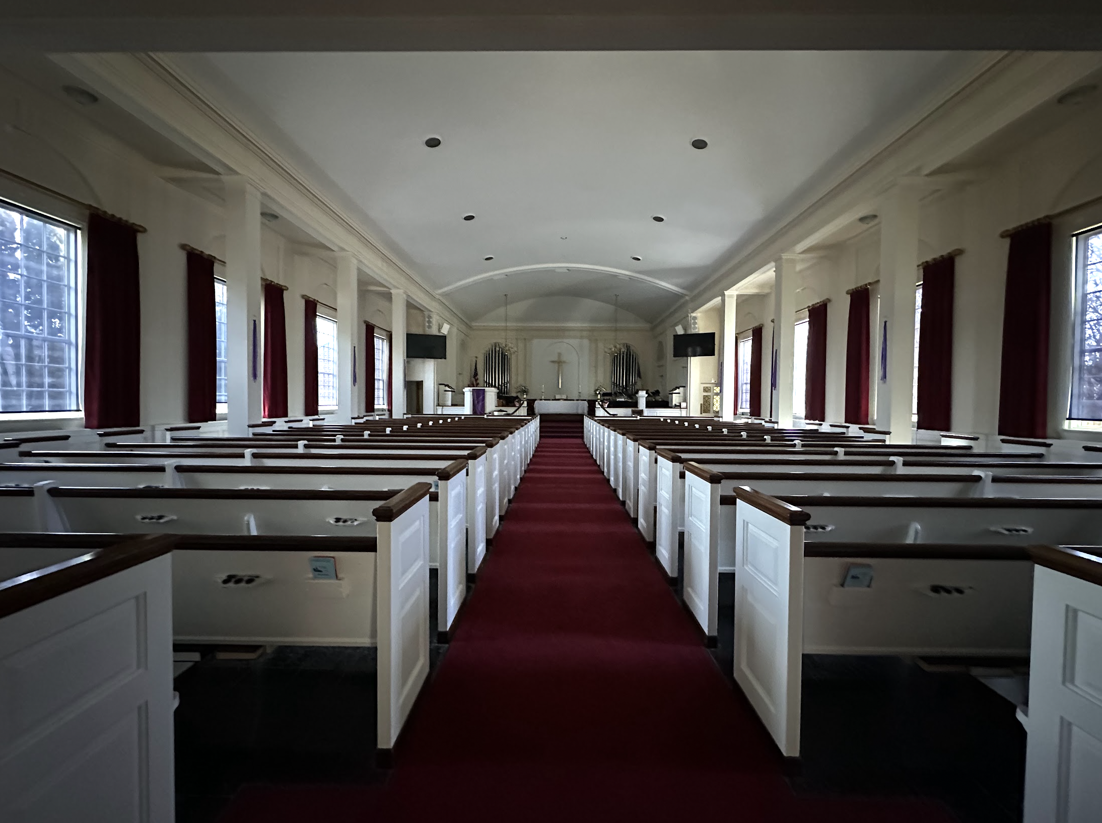 Interior of a church with white pews, a red aisle runner, altar at the front with cross, windows with red curtains, and organ pipes.