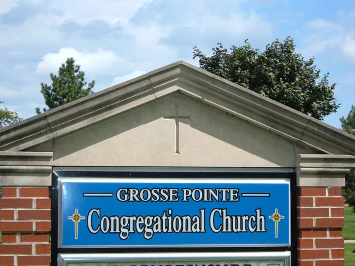 Sign for Grosse Pointe Congregational Church, with brick and stone building in the background, trees, and a partly cloudy sky.