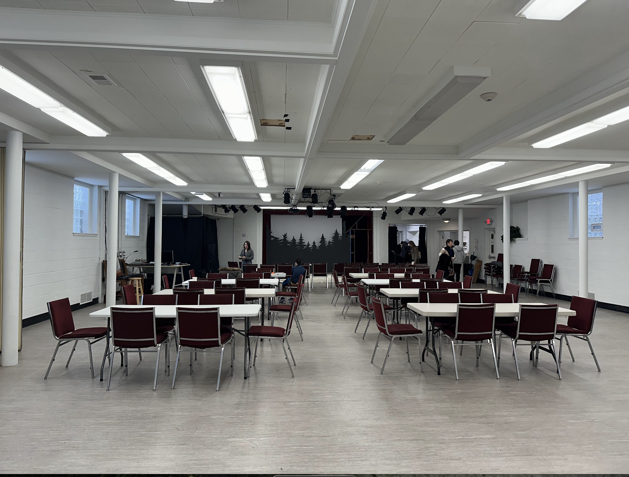 Empty multipurpose room with round tables and red chairs, a stage with a forest scene, and a few people preparing for an event.