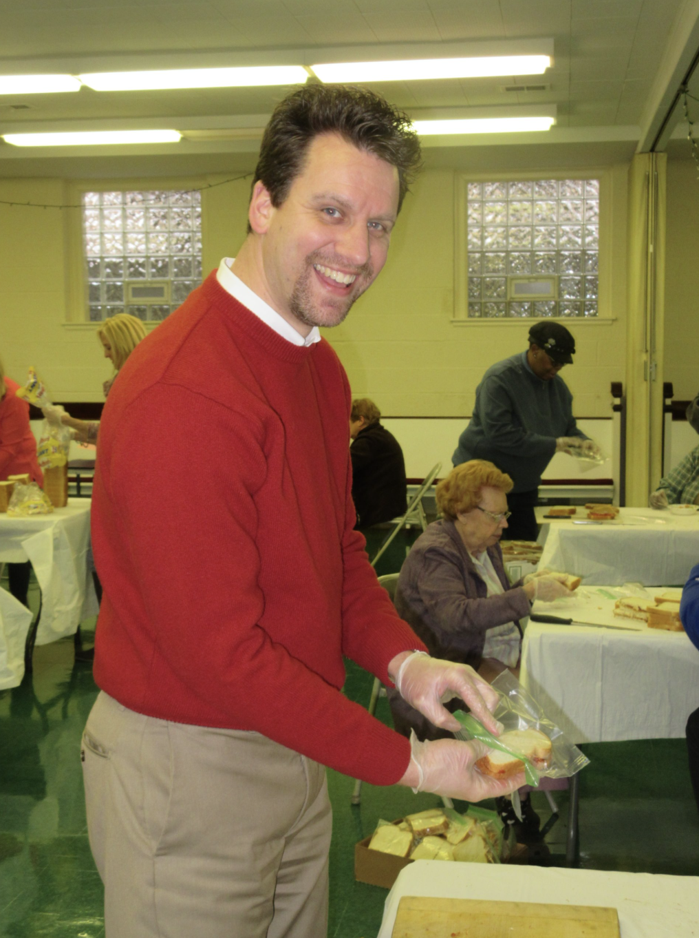 A man wearing a red sweater and gloves is smiling at the camera while preparing sandwiches at a community service event. In the background, women are working at tables with food in a large, well-lit room with yellow walls.