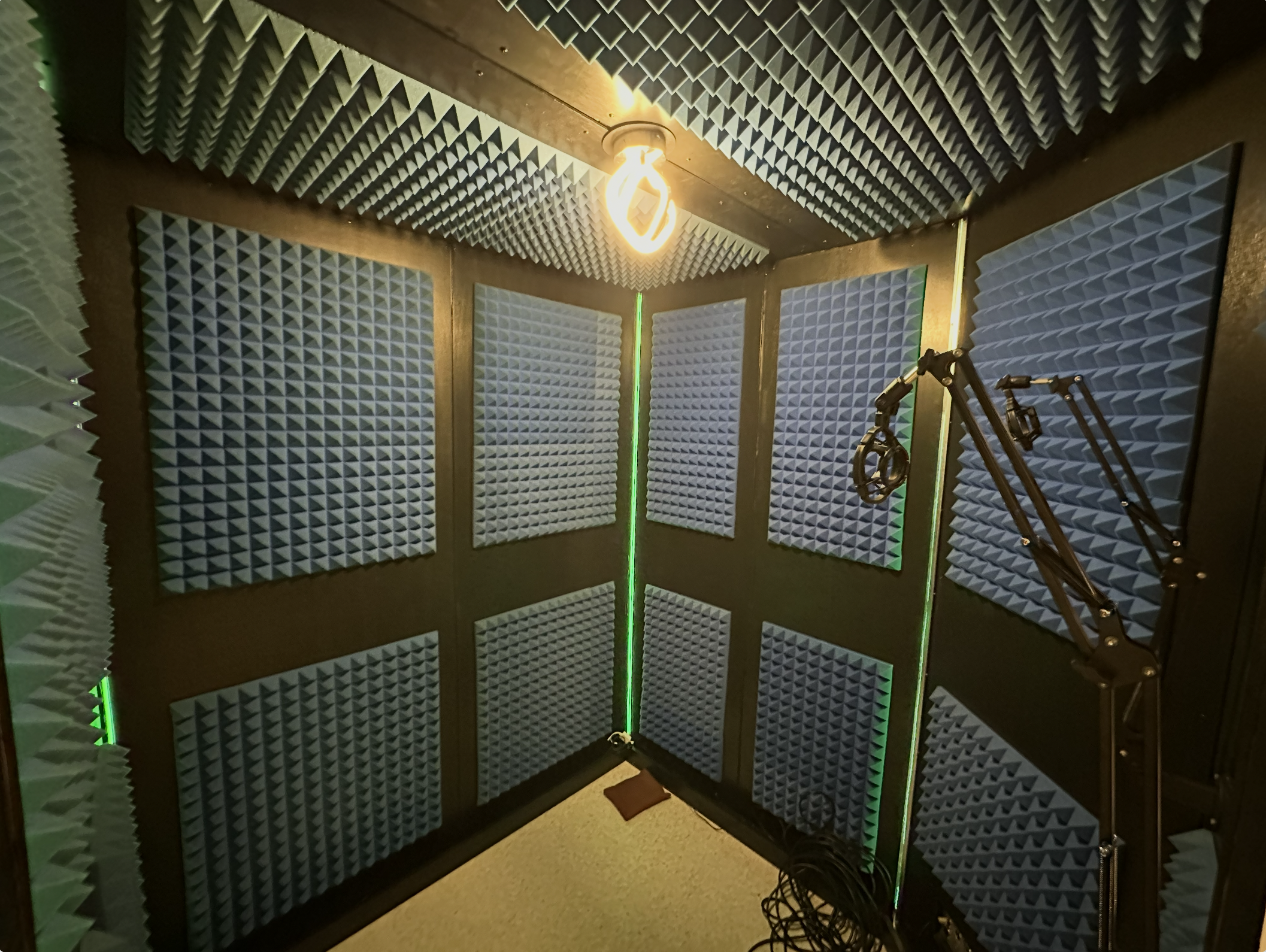 Small recording studio with black acoustic foam panels on the walls and ceiling. A bright lightbulb hangs from the ceiling, and a black microphone boom arm is attached in the corner, with cables on the floor.