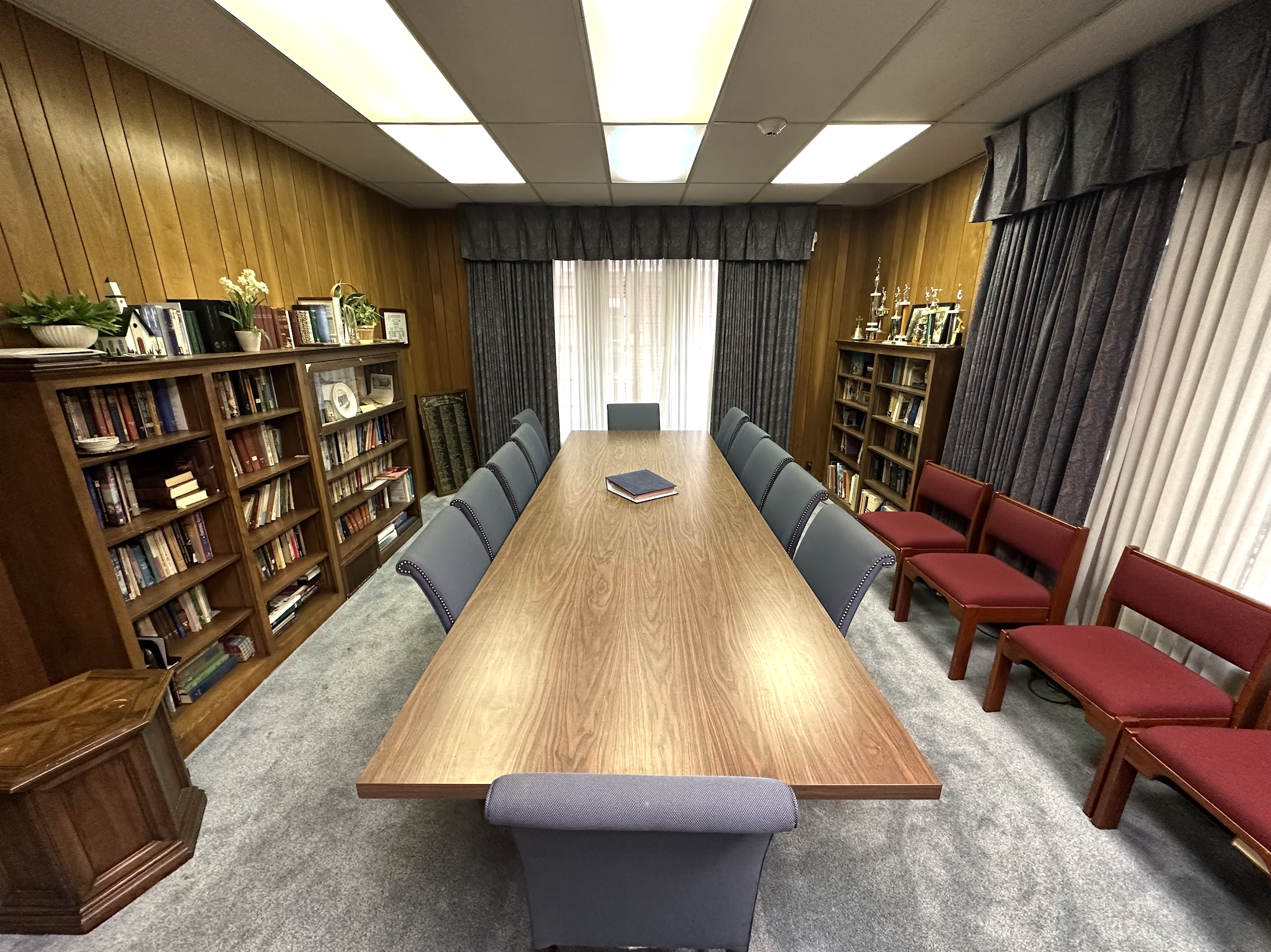 A conference room with a long wooden table surrounded by blue and red chairs, with bookshelves filled with books and trophies, and curtains covering windows.