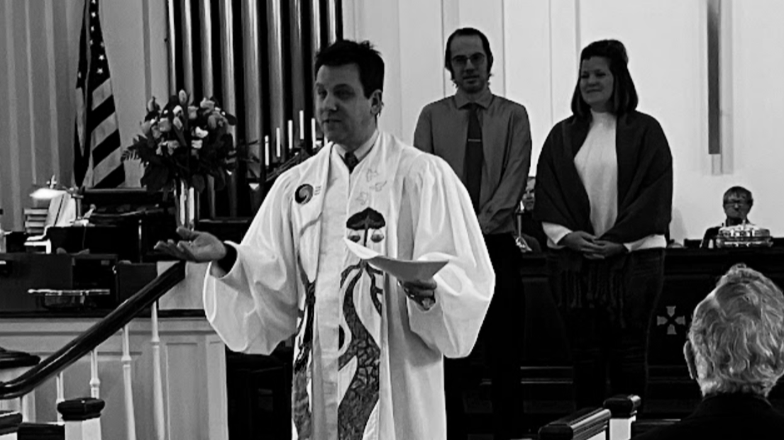 A male pastor in a white robe, standing in front of a group, speaking, with two people standing behind him in an indoor setting church sanctuary.