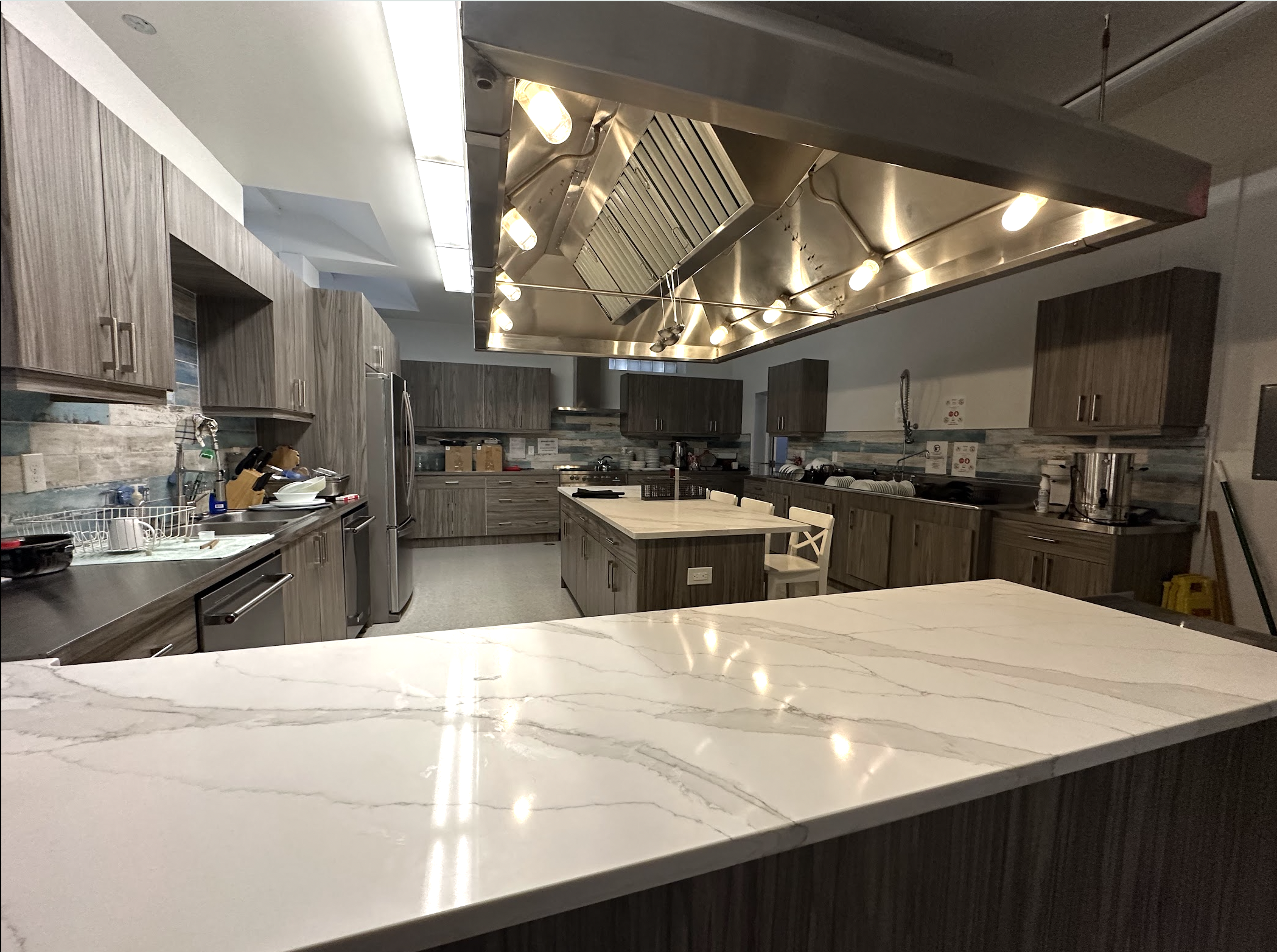 Modern kitchen with wooden cabinets, stainless steel appliances, marble countertops, and a large central island with a marble surface. Includes pendant lighting and various kitchen tools and supplies.