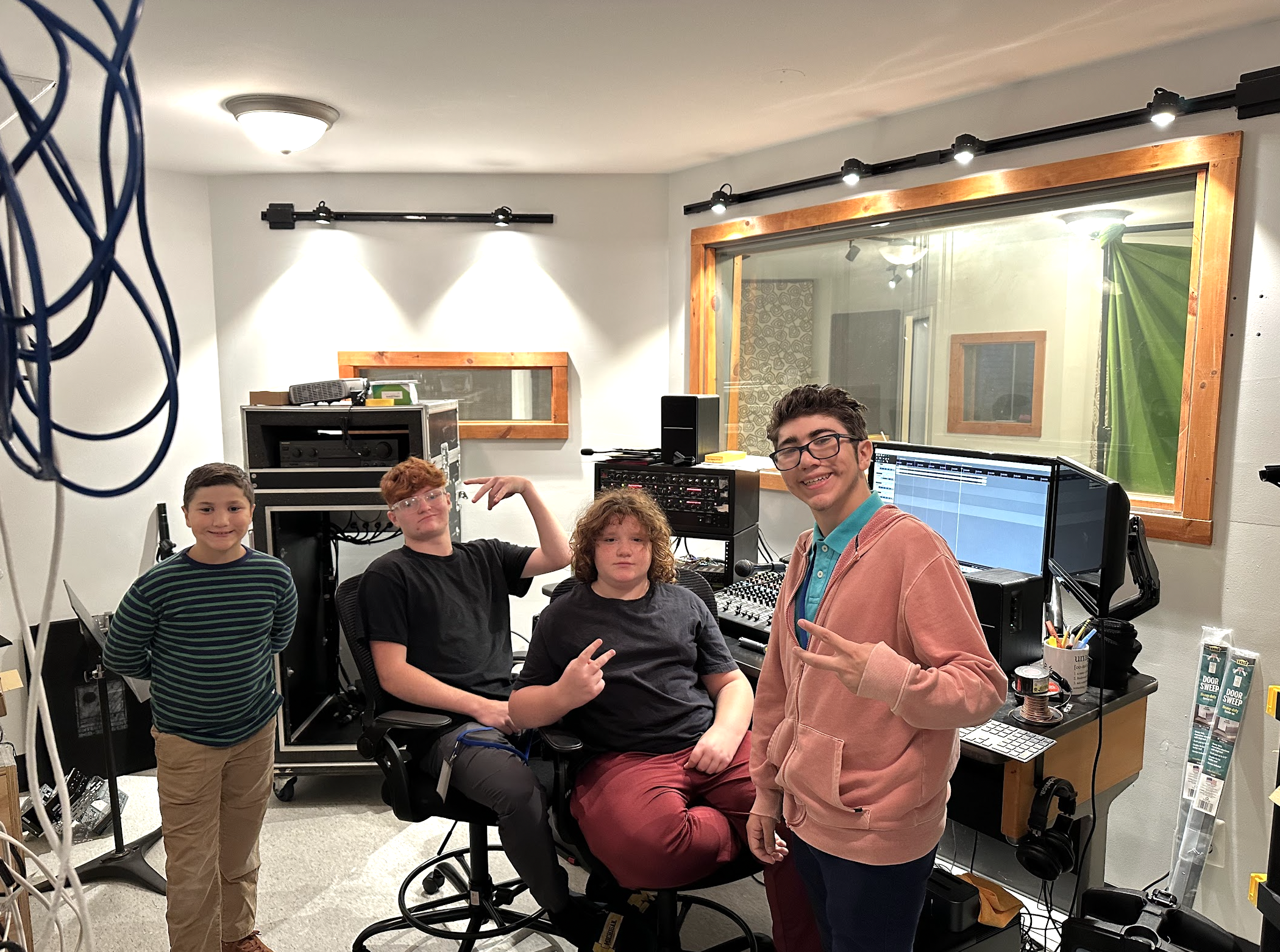 Four boys in a recording studio, some making peace signs, with audio equipment and a large window behind them.