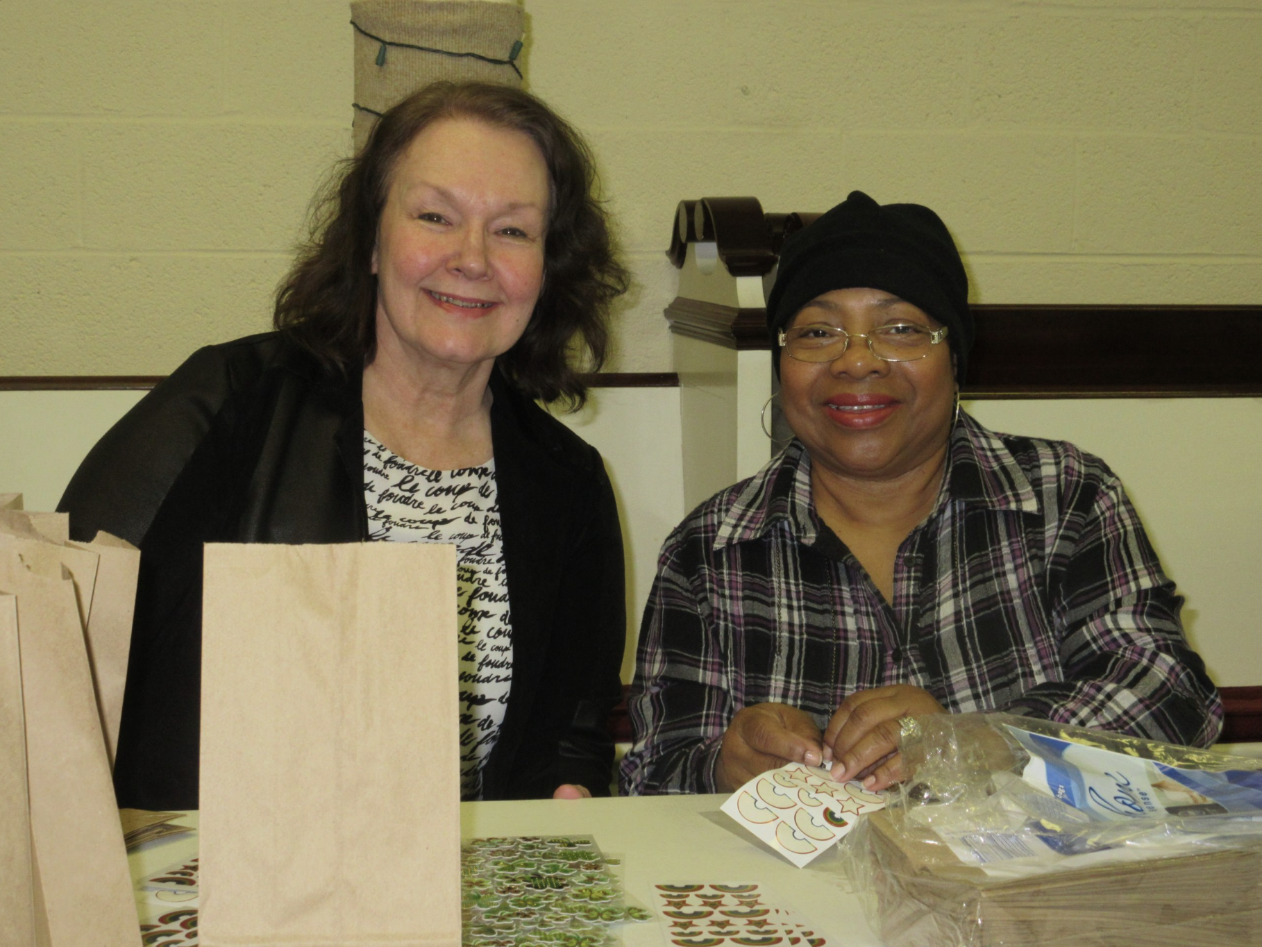 Two women sitting at a table, smiling. The table has stickers, paper bags, and a plastic-wrapped item. The woman on the left has shoulder-length dark hair, fair skin, and is wearing a black jacket and a white top with black handwritten script. The wo