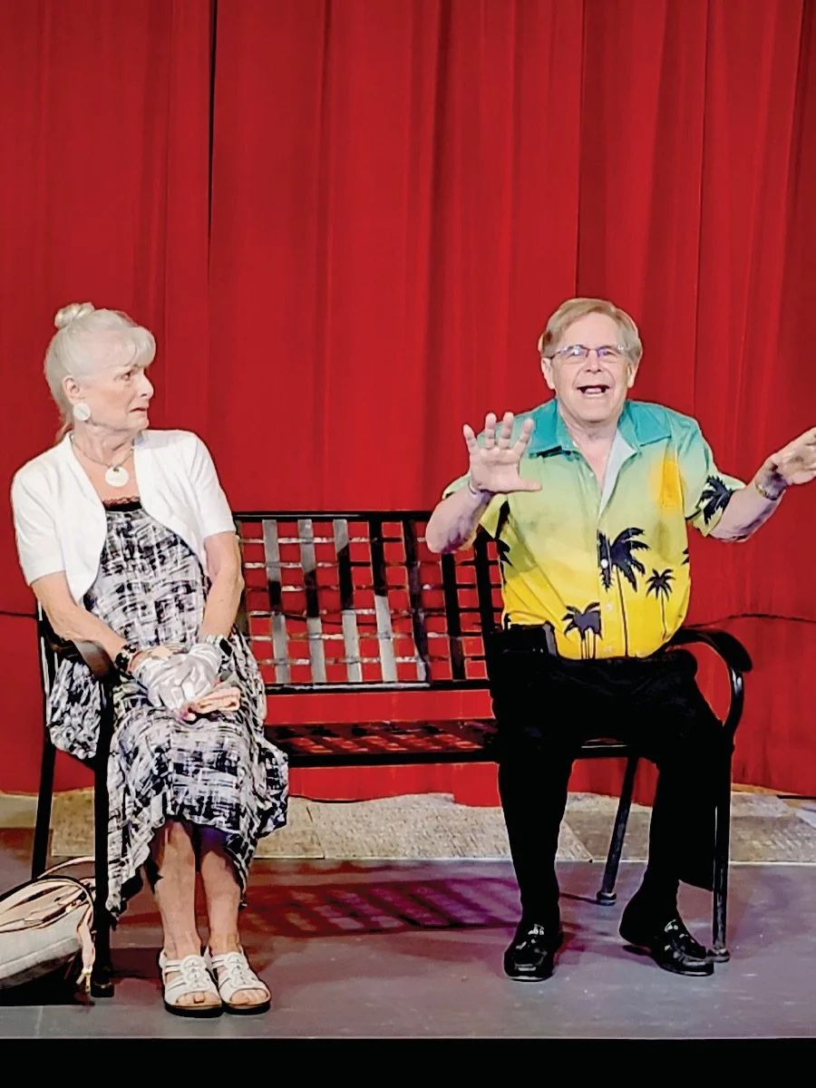 An elderly woman and a man sitting on a bench in front of a red curtain during a theater performance.  The woman is wearing a black and white patterned dress with a white cardigan, and the man is wearing a colorful Hawaiian shirt with palm trees. 