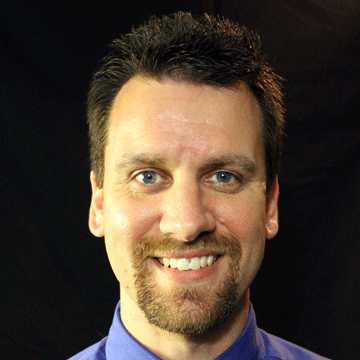 A smiling man with short dark hair, blue eyes, and a beard, wearing a blue shirt against a dark background.