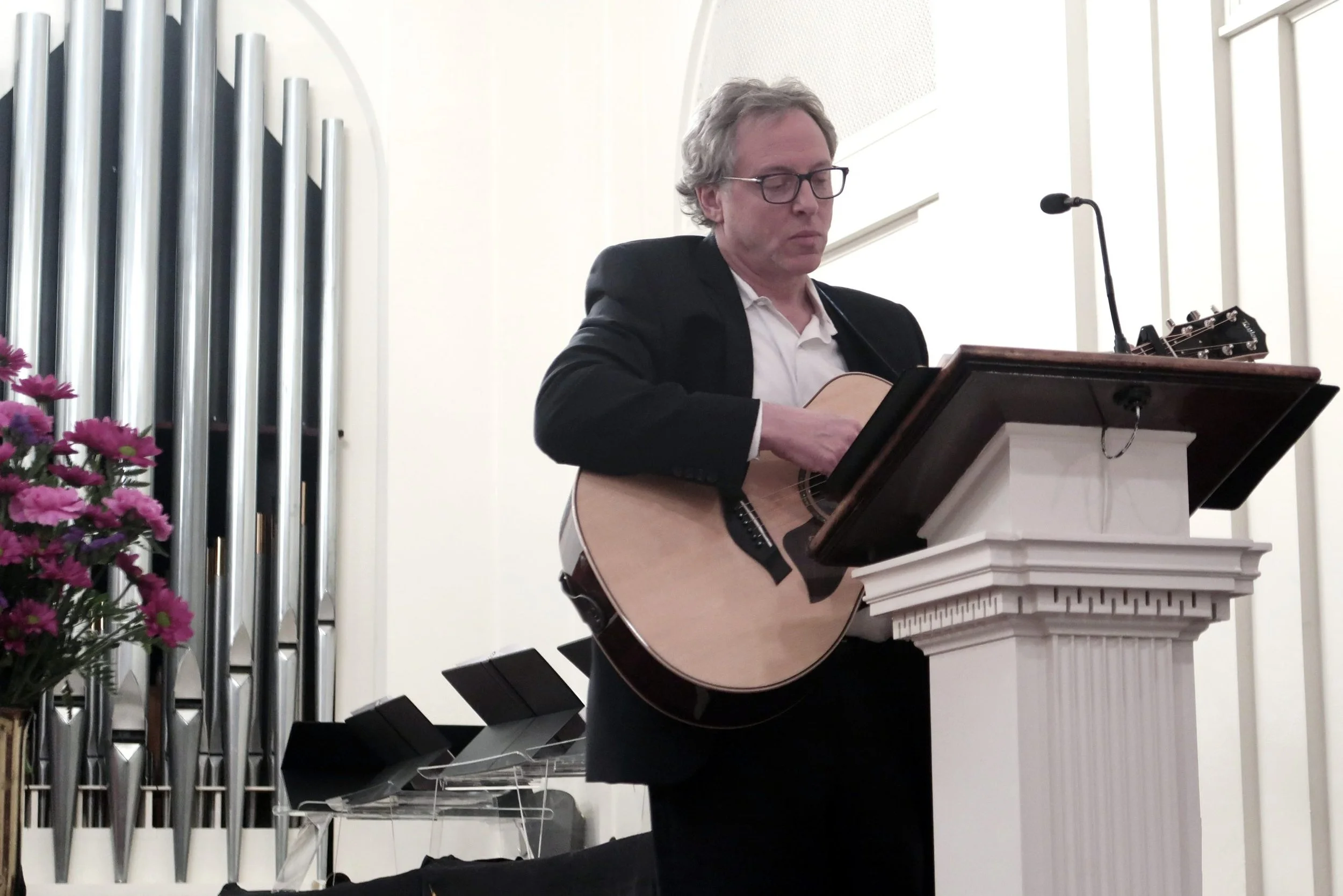 Man playing an acoustic guitar while standing at a music stand in a church or chapel.