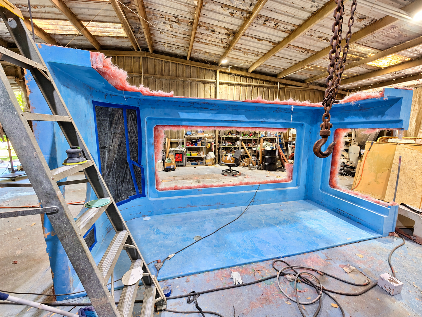 Interior of a large blue foam structure under construction inside a workshop, with a ladder on the left side, work tools and supplies scattered around, and shelves with various materials in the background.