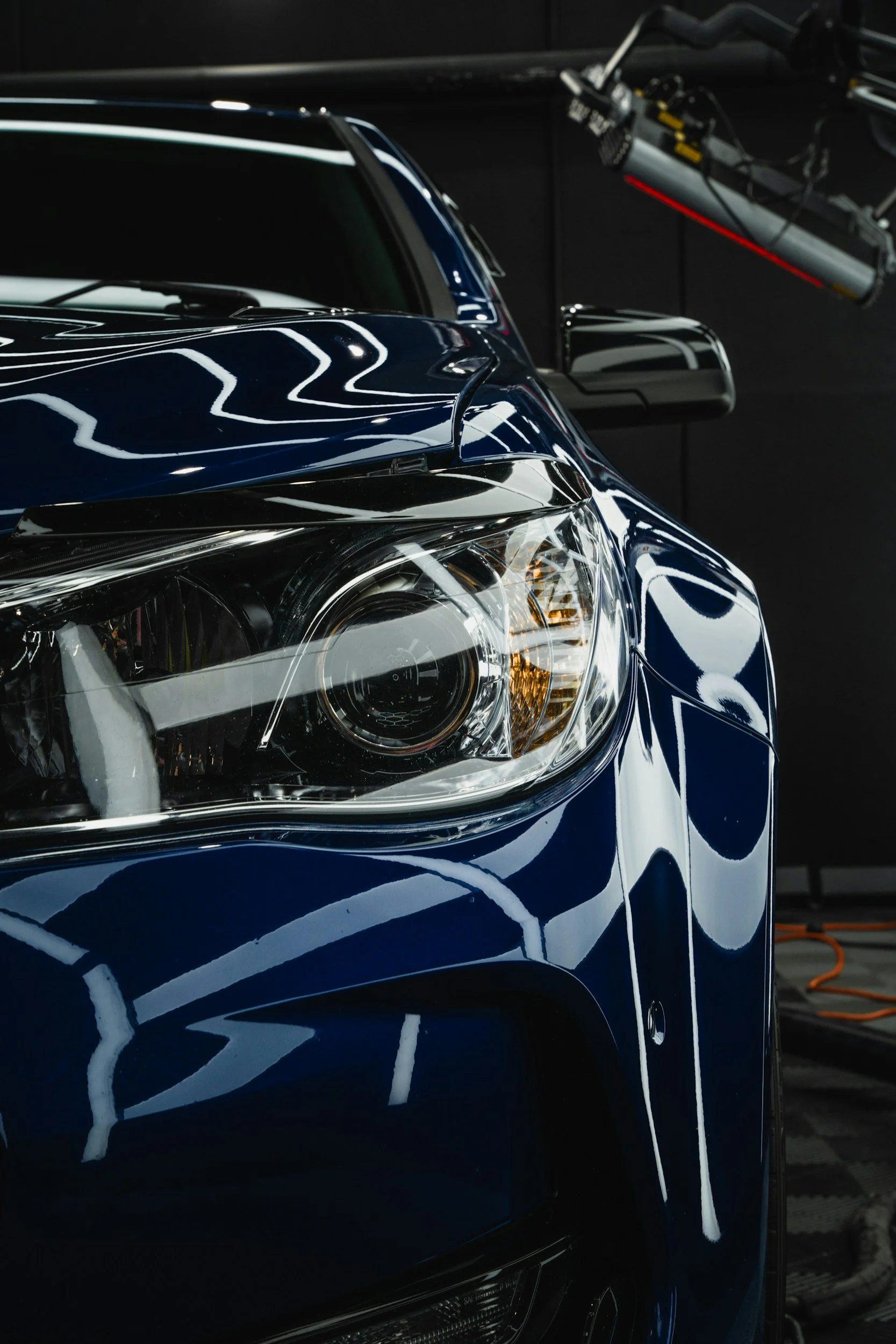 Close-up of the front headlight and hood of a shiny blue car in a garage, with reflections on its surface and a garage light above.