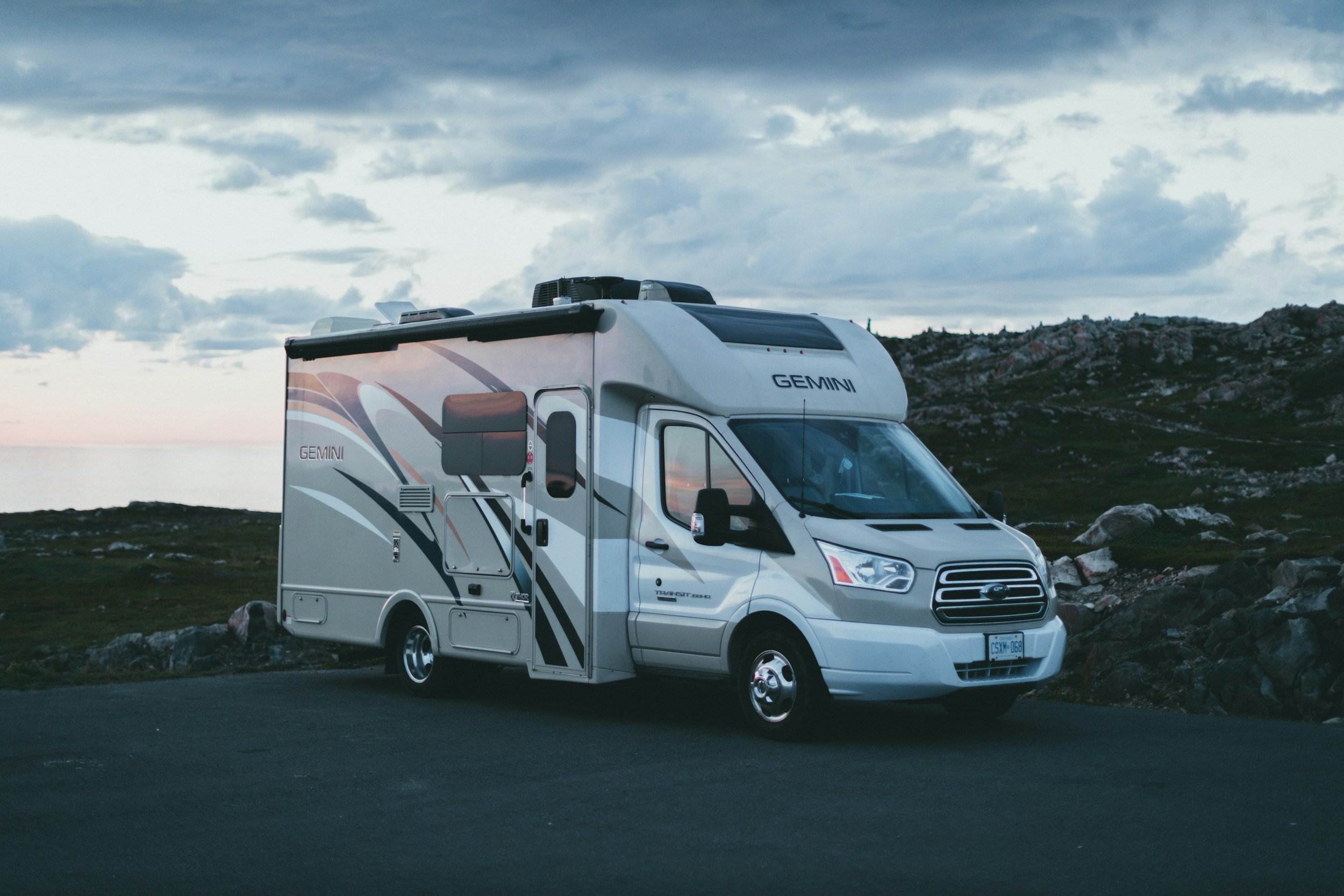 A white and gray camper van labeled 'GEMINI' parked on a paved road near rocky terrain with a body of water and cloudy sky in the background.