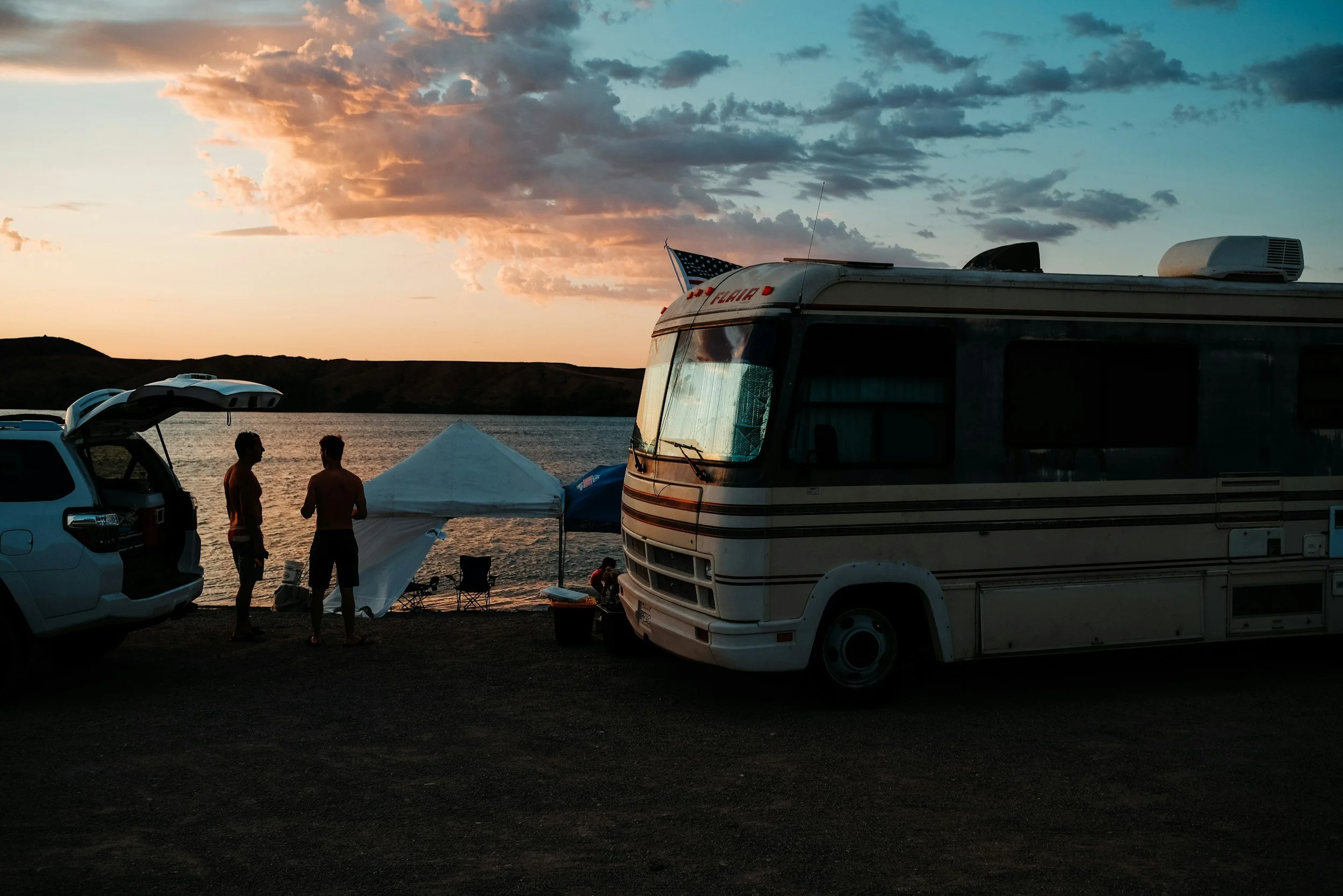Two people stand near a car and a camper van by a lake at sunset, with a small white tent and a couple of chairs, during a camping trip.