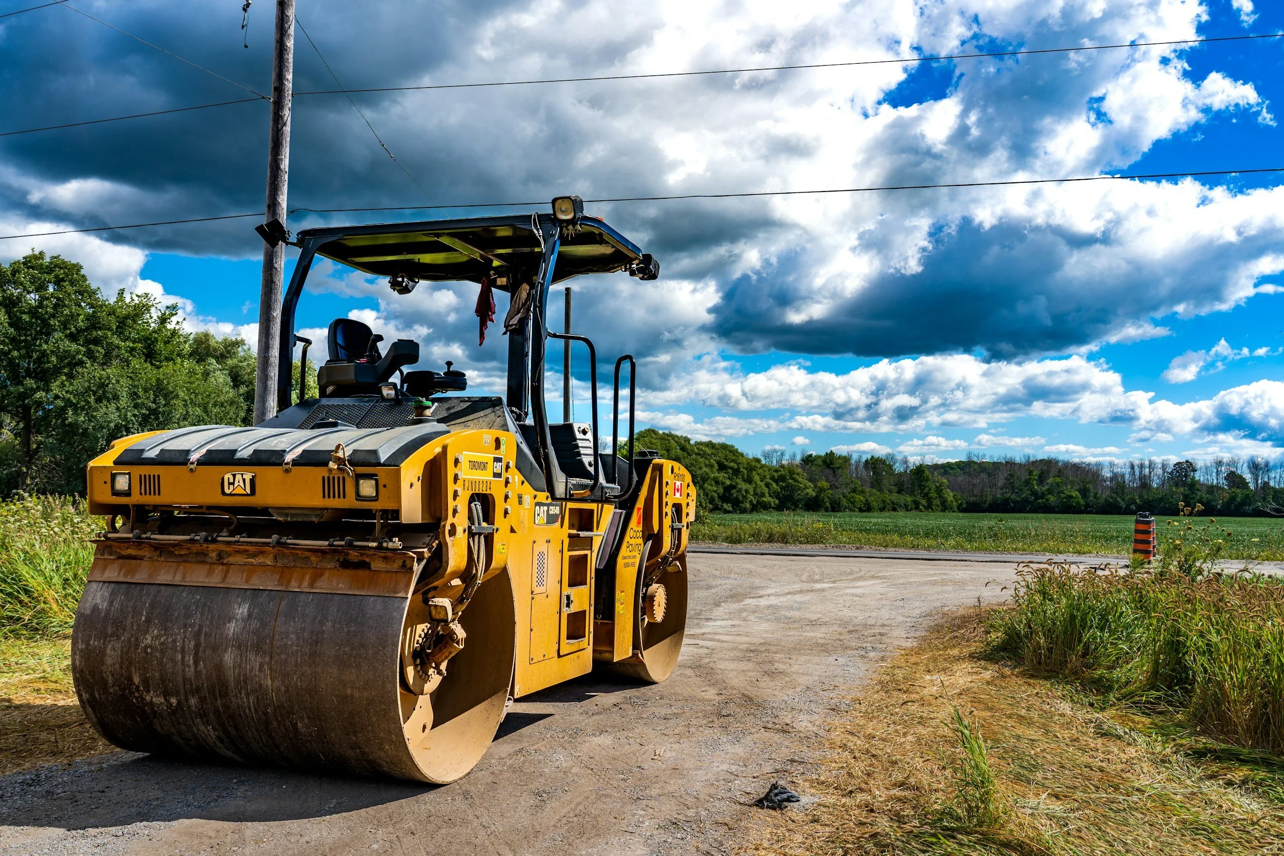 A yellow CAT steamroller on a construction site with a dirt road, green trees, and a cloudy blue sky in the background.