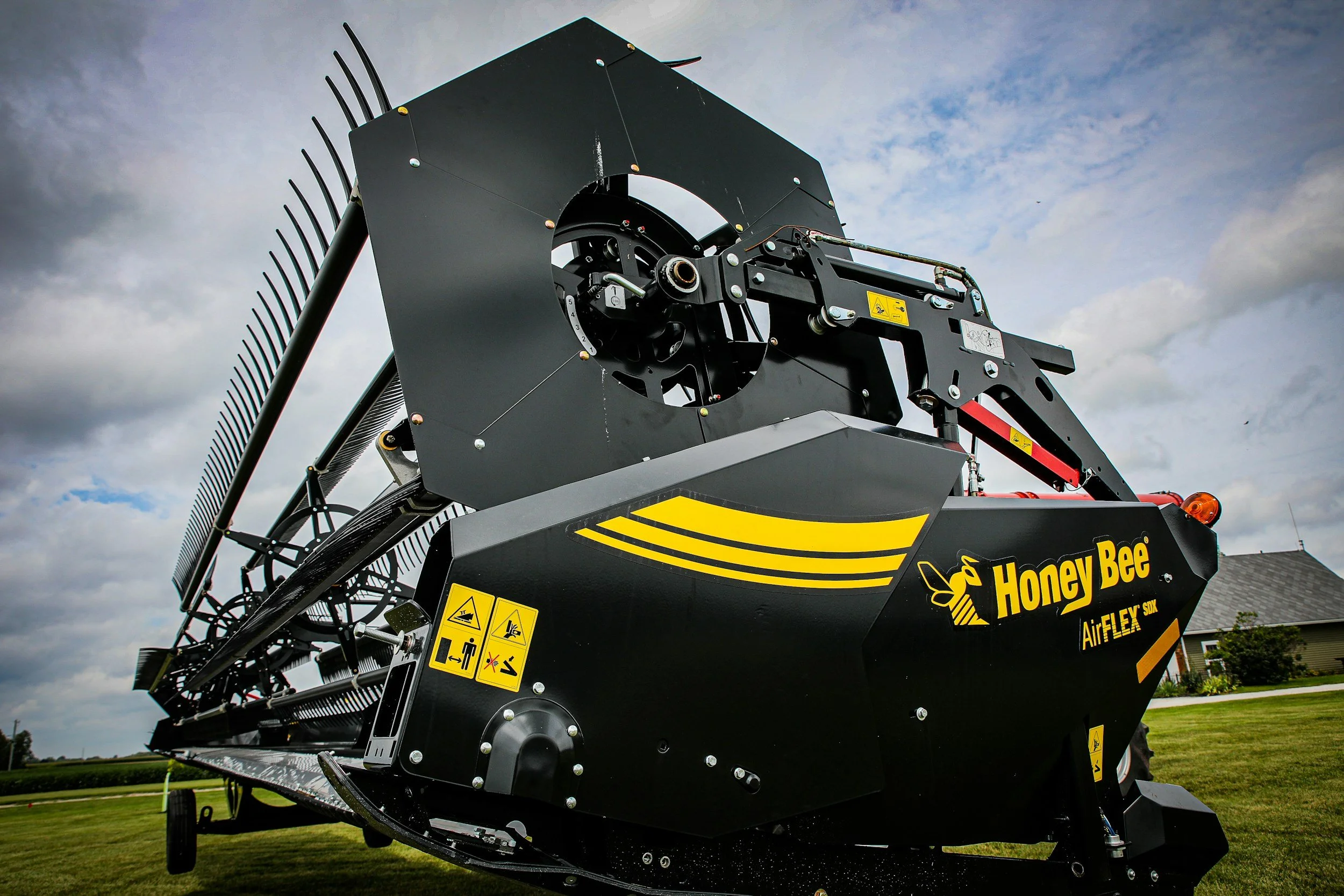 A large black and yellow Honey Bee agricultural machine, possibly a hay mower or harvester, situated on a grassy field with a partly cloudy sky in the background.