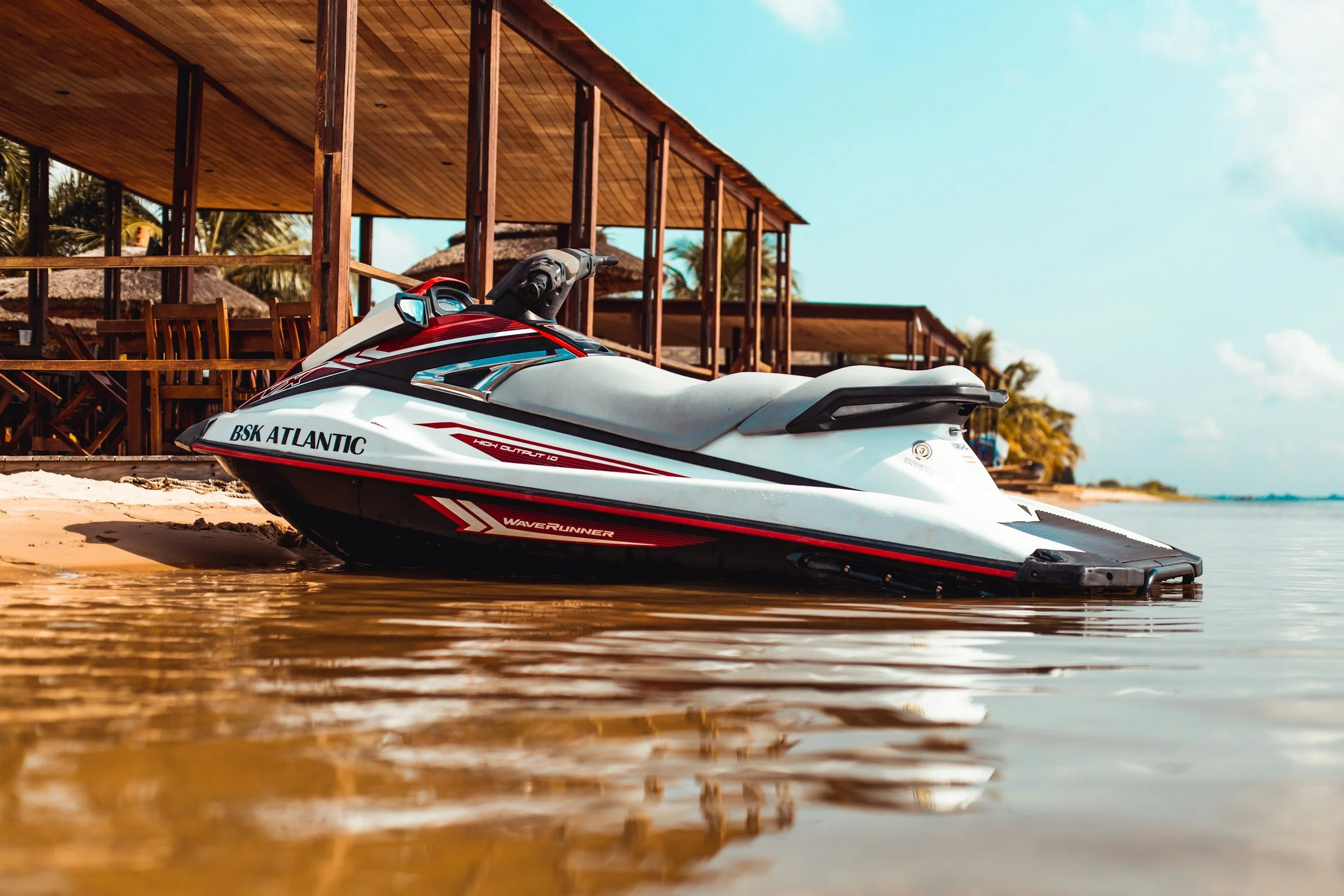 A white and black Yamaha Waverunner jet ski with red accents resting on the shore of a beach near a wooden resort structure, with palm trees and a blue sky in the background.