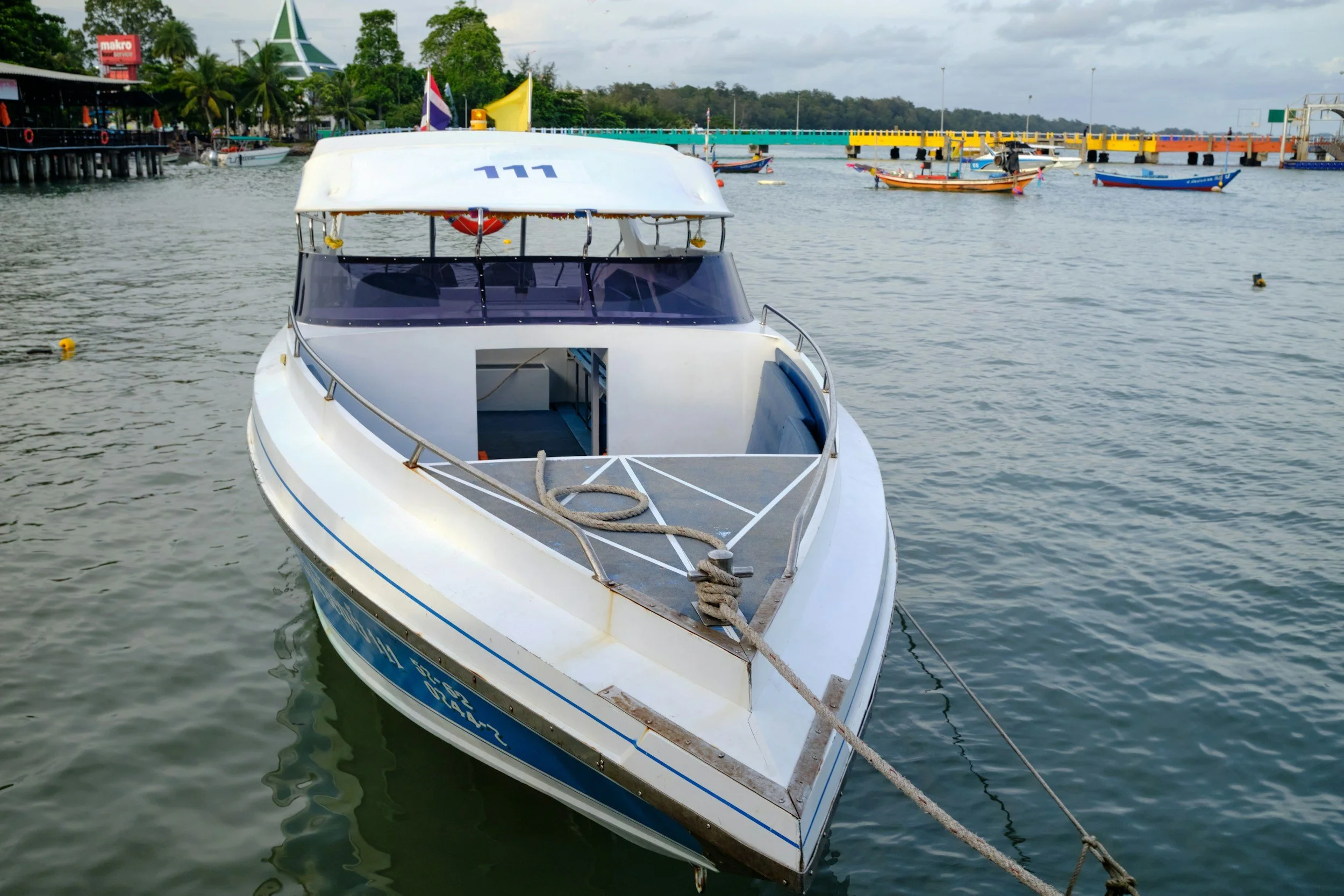 A white motorboat docked in a harbor with several smaller boats in the background, a bridge, and a recreational area with greenery and buildings.