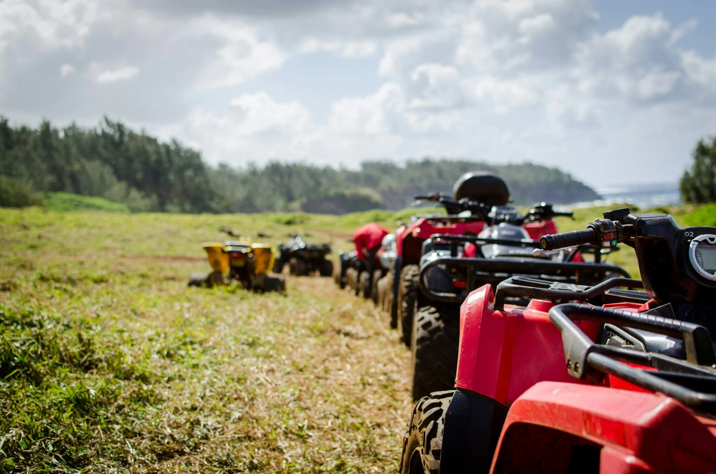Line of all-terrain vehicles (ATVs) parked on grassy field under partly cloudy sky with trees in background
