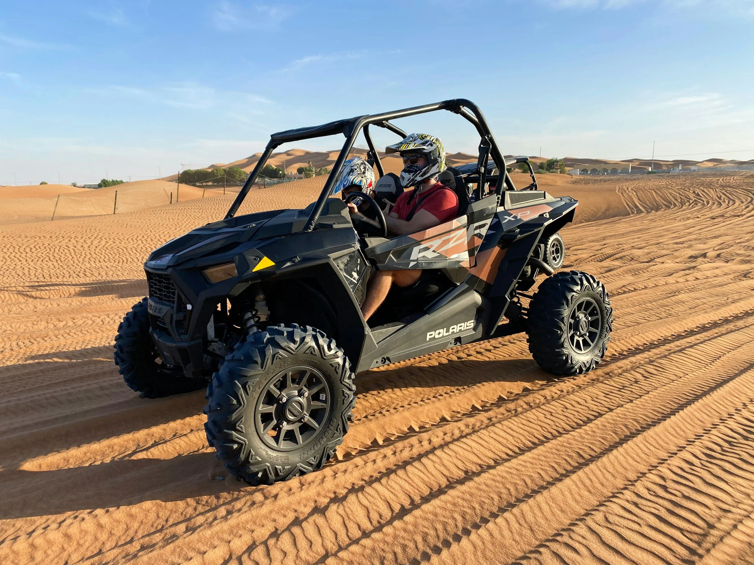 A black Polaris off-road vehicle with two riders wearing helmets is driving across sandy desert terrain with sand dunes in the background.