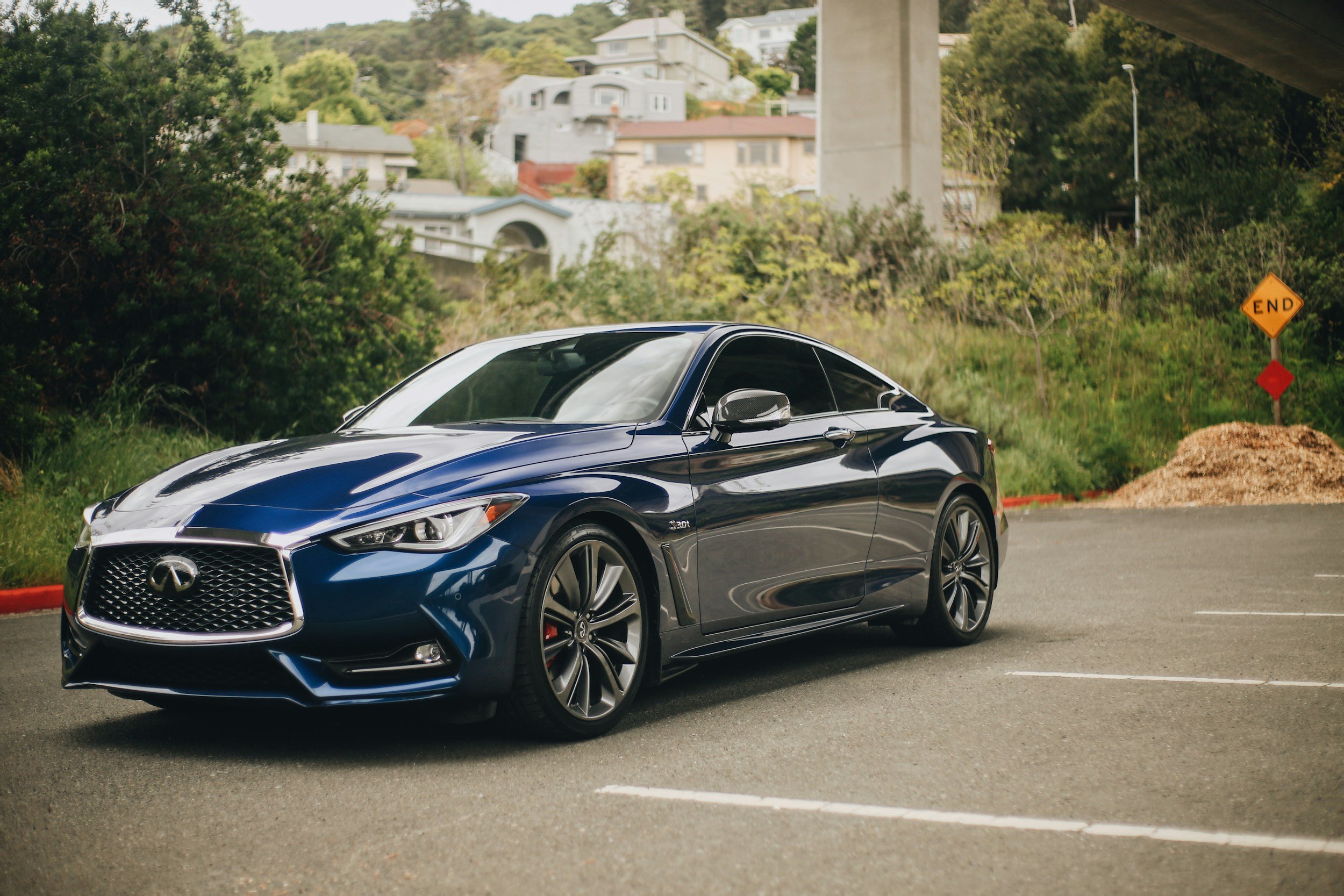 A dark blue Infiniti sports car parked in an empty parking lot with greenery and houses in the background.