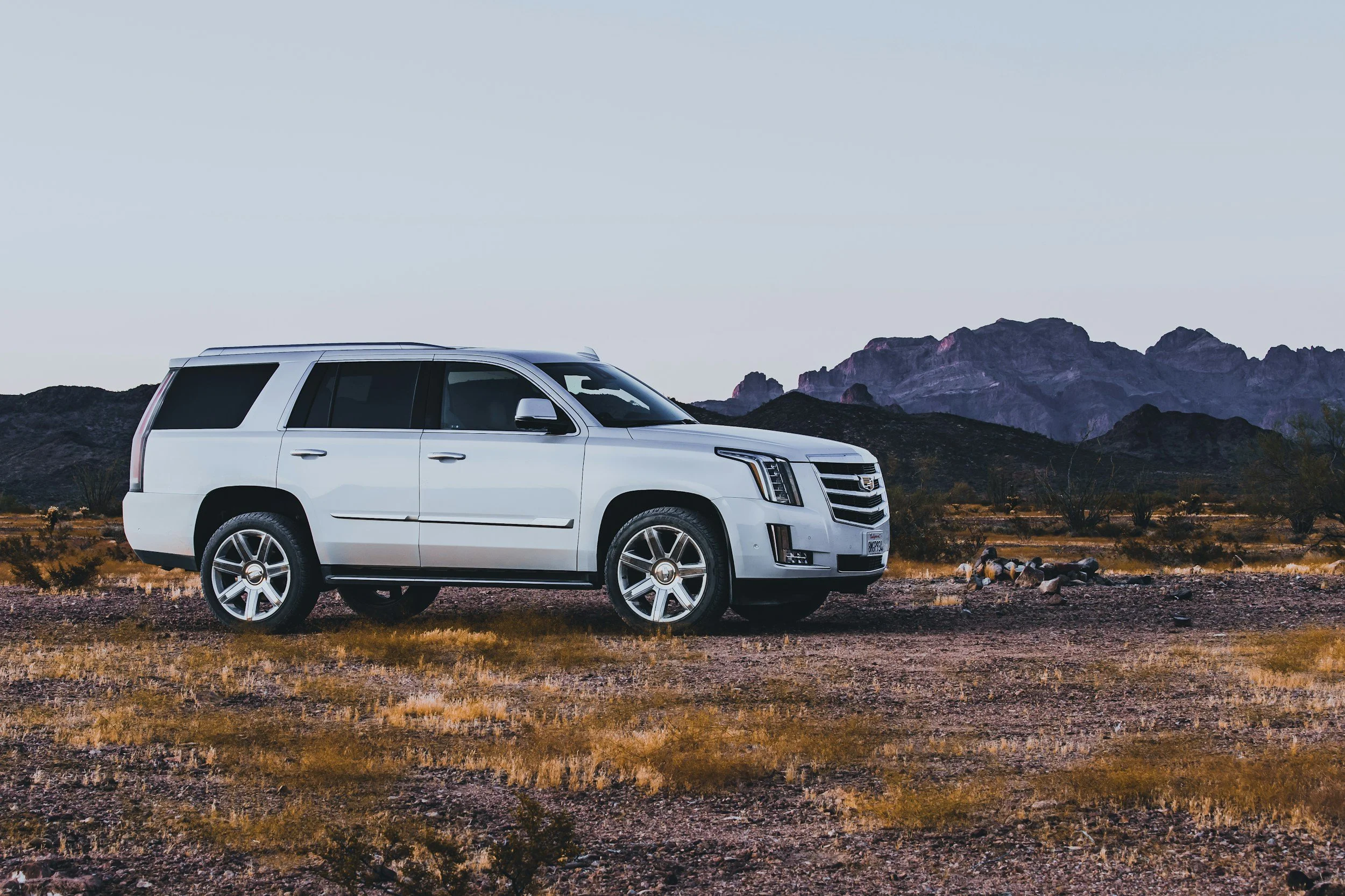 A white Cadillac SUV parked on a rocky desert landscape with sparse vegetation, mountains in the background, and a clear sky.