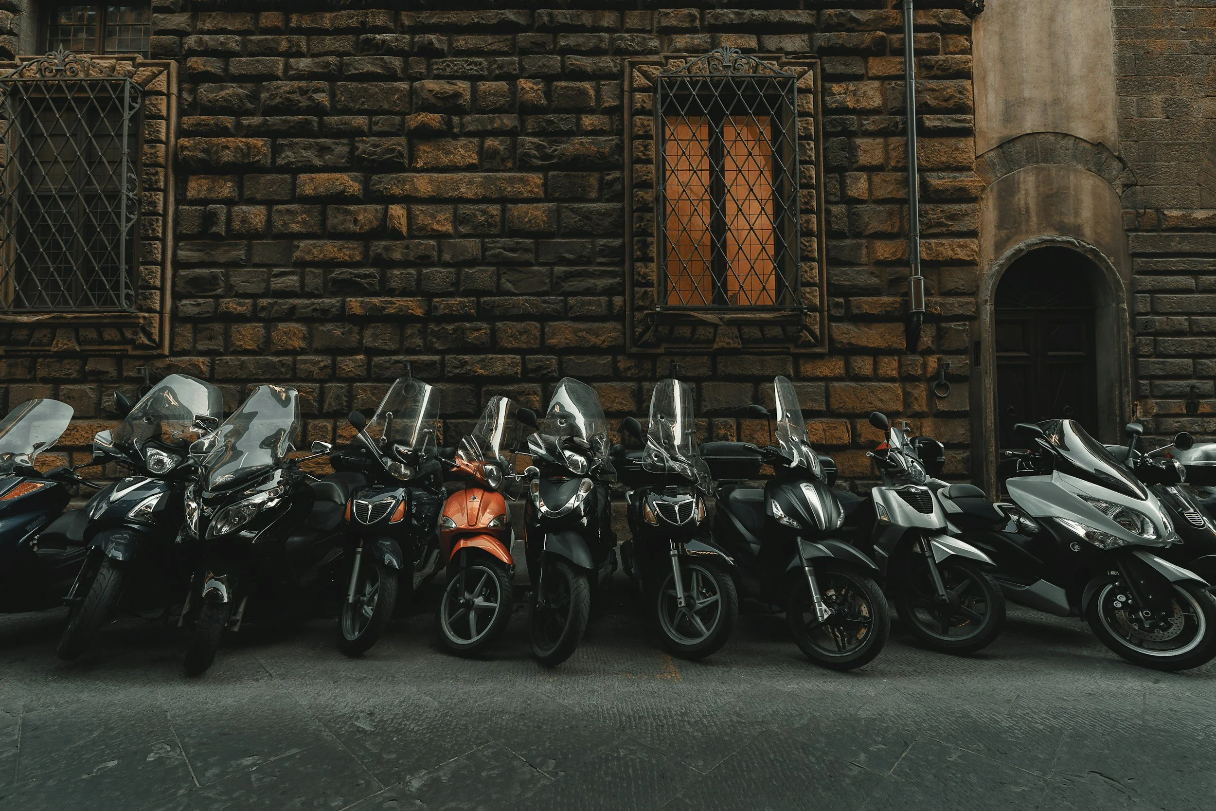 Multiple motorcycles and scooters parked in front of a brick building with barred windows and an arched doorway.