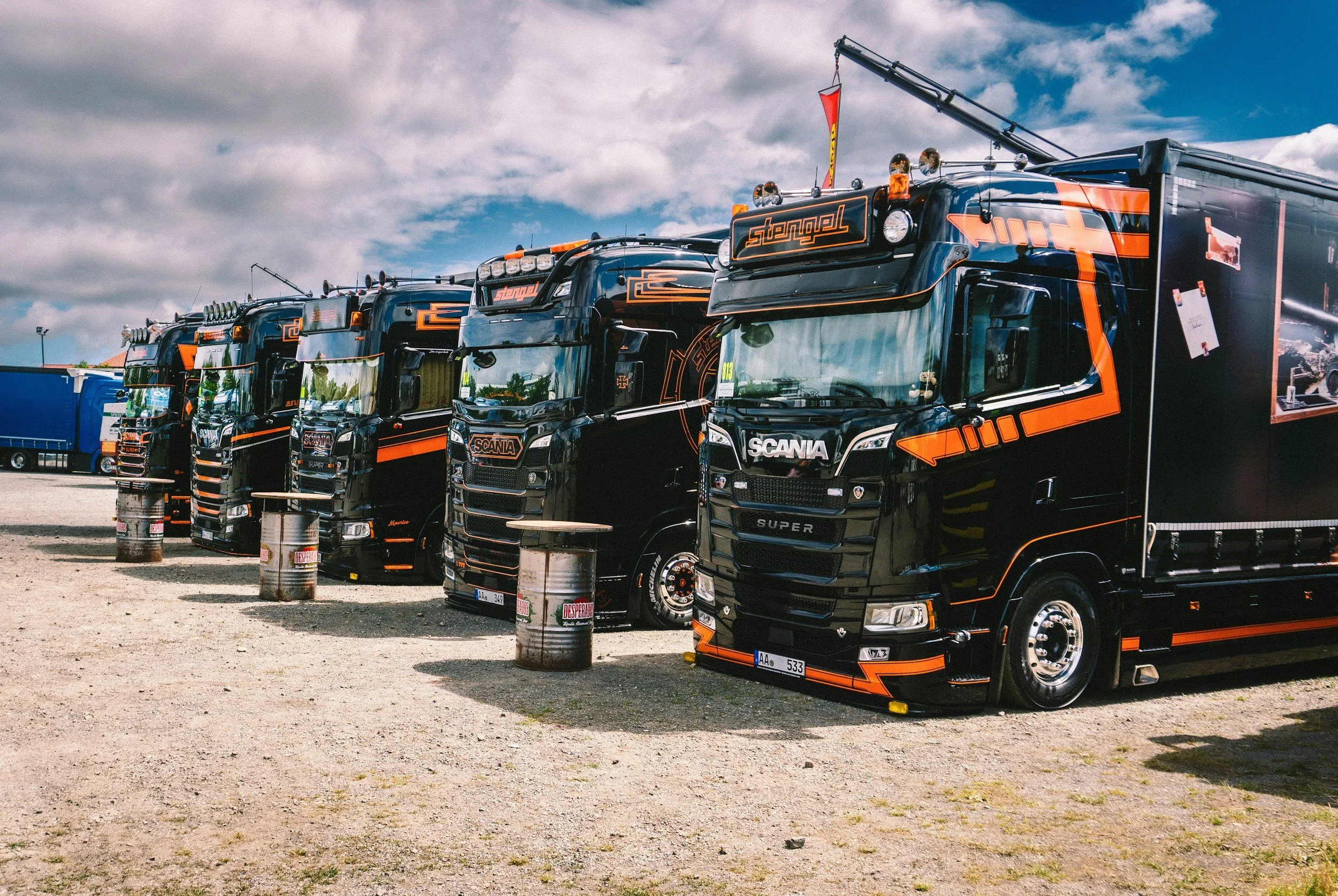 A row of black racing trucks with orange accents parked outdoors on a gravel surface under a partly cloudy sky, with some barrels in front of each truck.