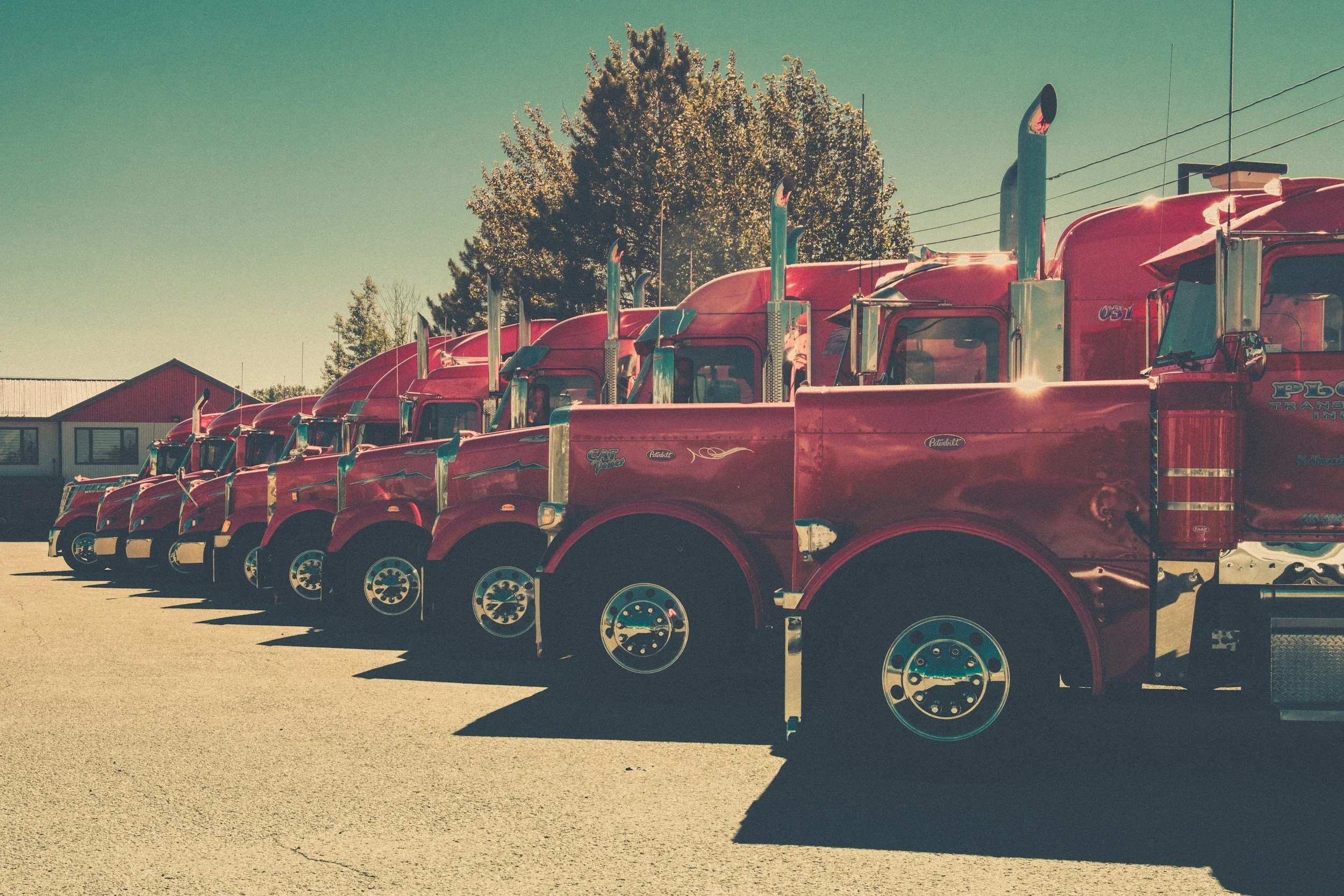 A row of shiny red semi-trucks parked side by side in a lot with shadows cast on the ground, under a clear sky with trees and a building in the background.