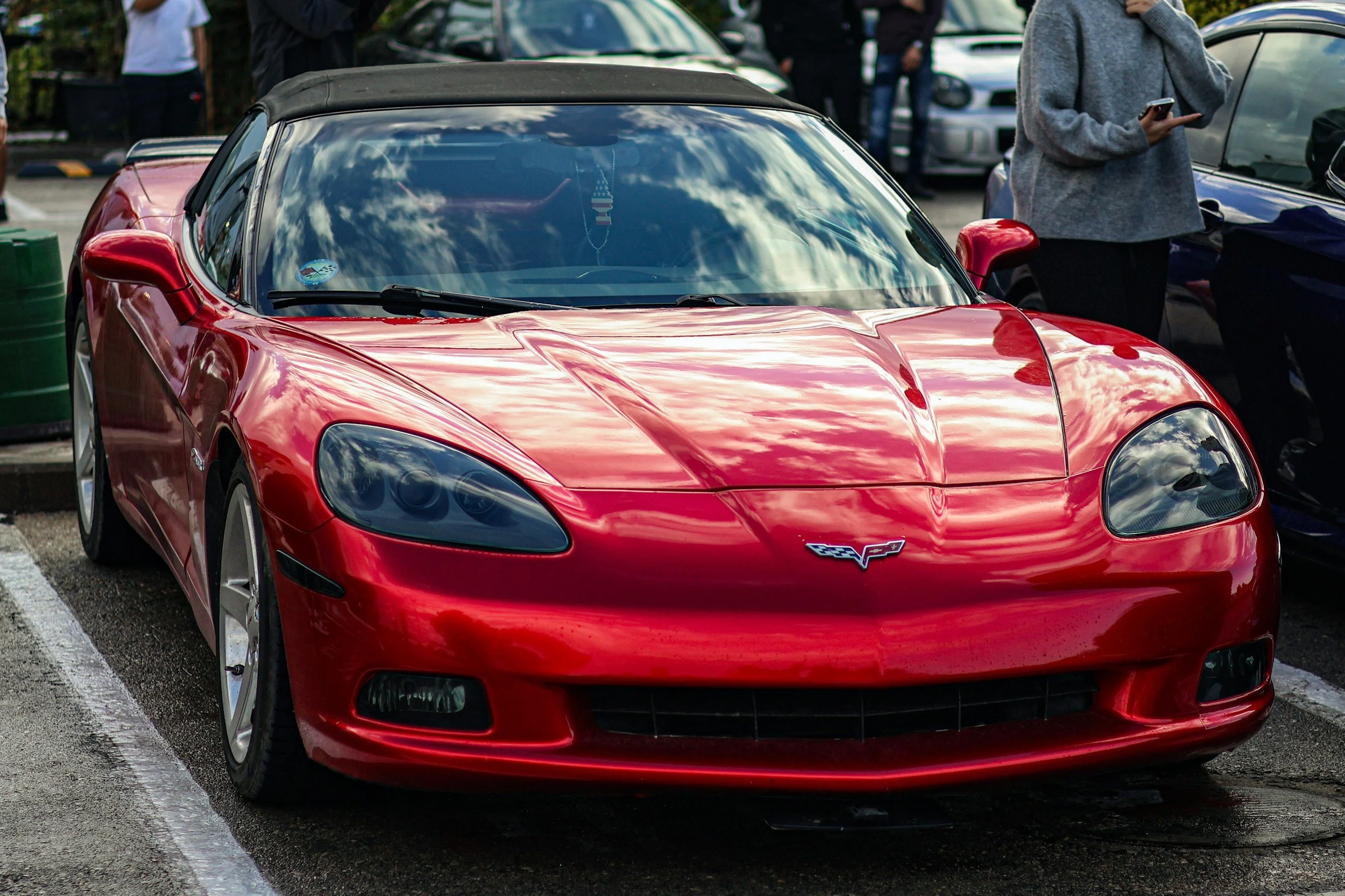 Red Chevrolet Corvette sports car parked in a lot, with people standing around and a cloudy sky reflected on the windshield.