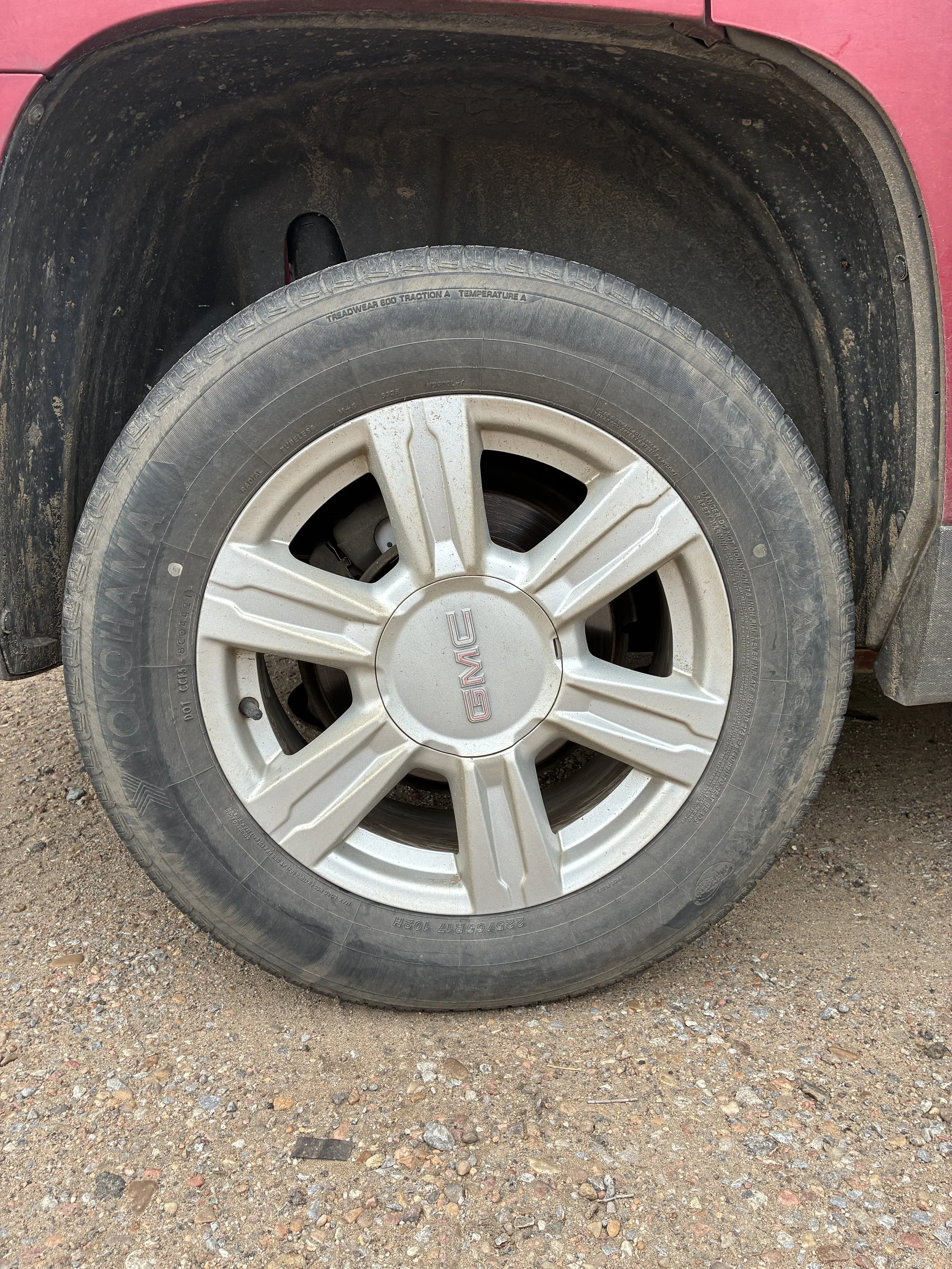 A close-up photo of a car tire with a silver alloy rim, mounted on a red vehicle. The tire is dirty, and the car's wheel well is also dusty and dark.
